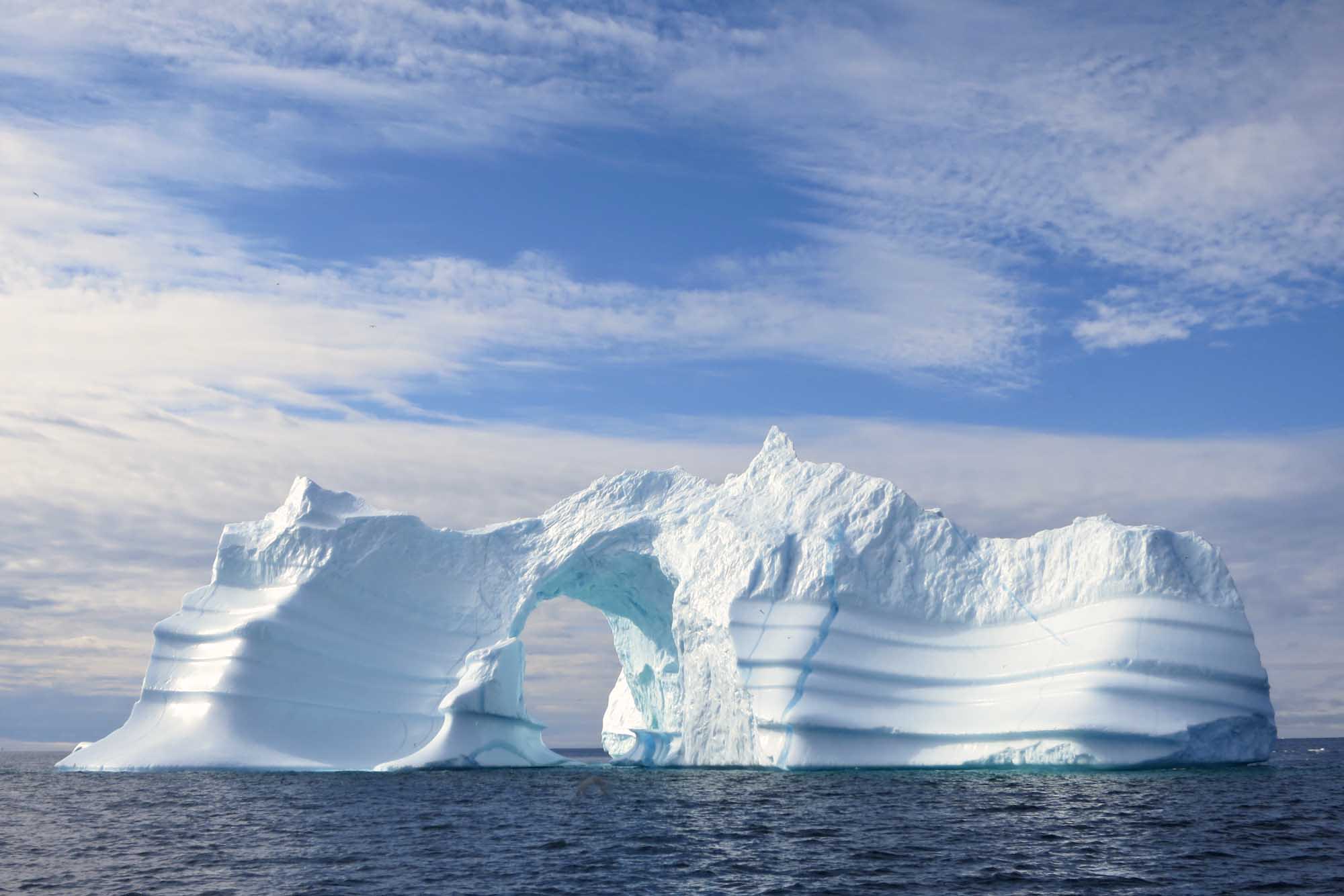 A striking natural arch iceberg floats in the dark waters of Greenland, featuring brilliant white
