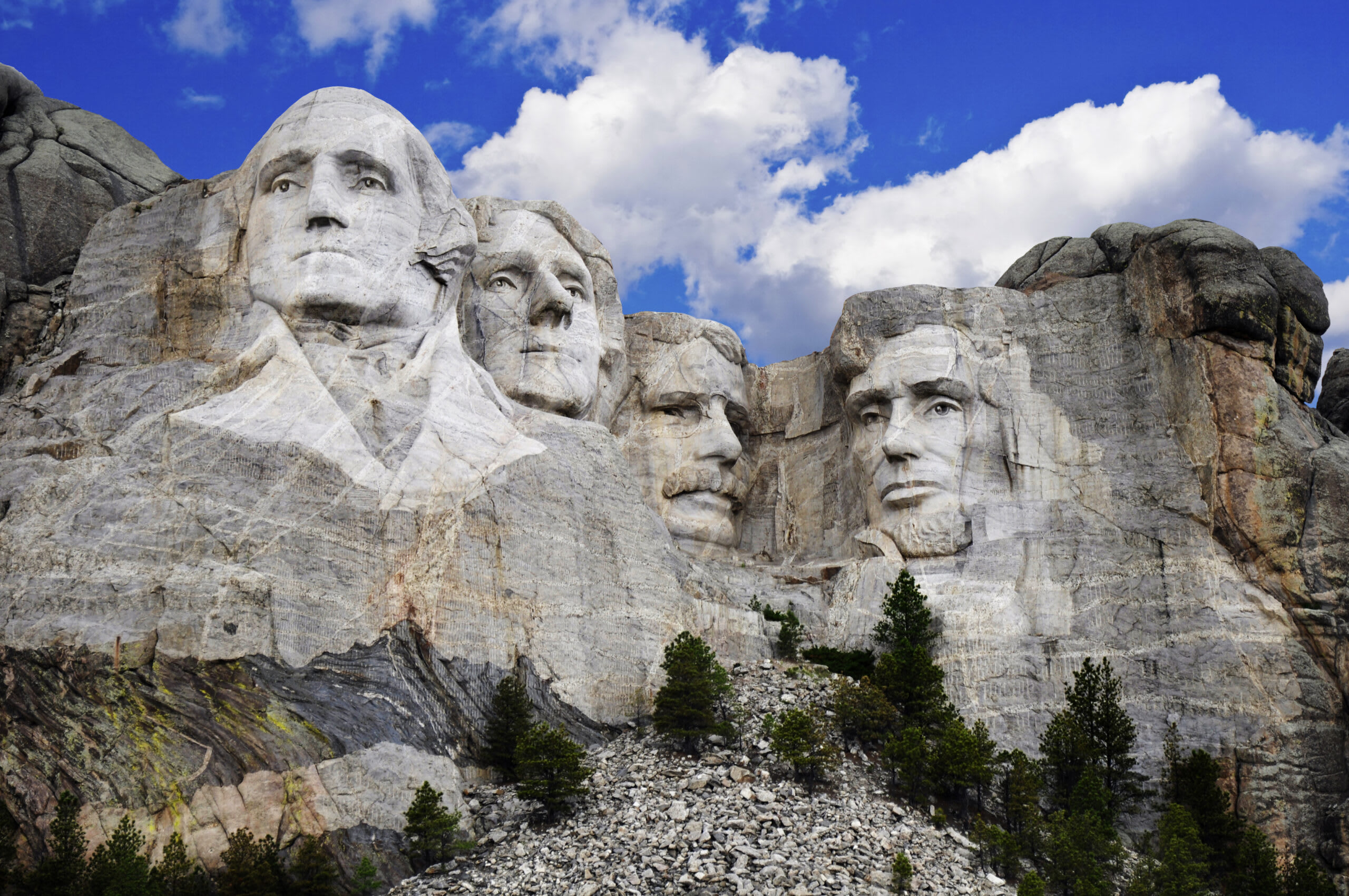 The iconic Mount Rushmore monument featuring four carved presidential faces (Washington, Jefferson, Theodore Roosevelt, and Lincoln) carved into granite cliffs against a brilliant blue sky with white clouds. Evergreen trees dot the base of the mountain.