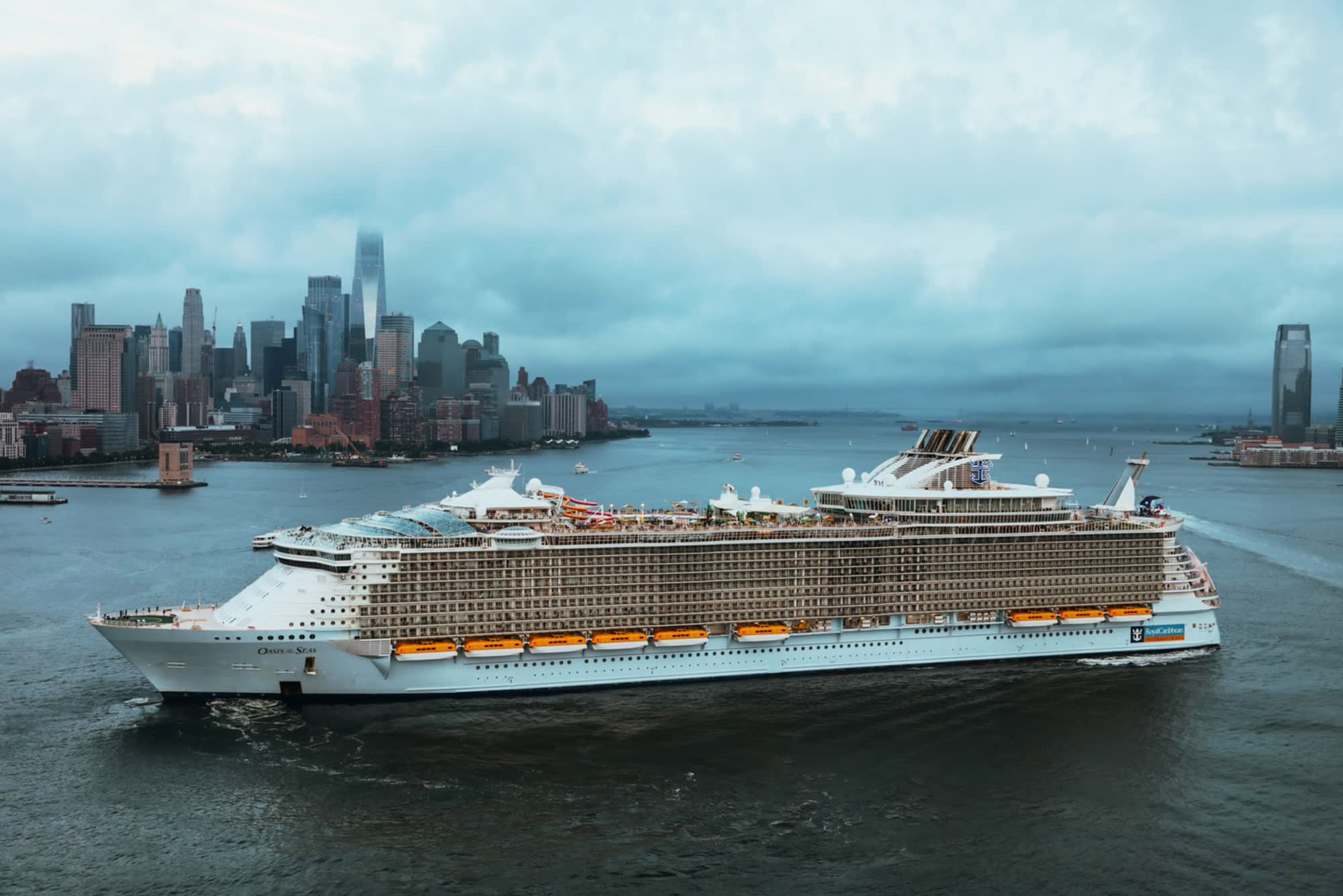 A massive modern cruise ship anchored in New York Harbor with the Manhattan skyline in