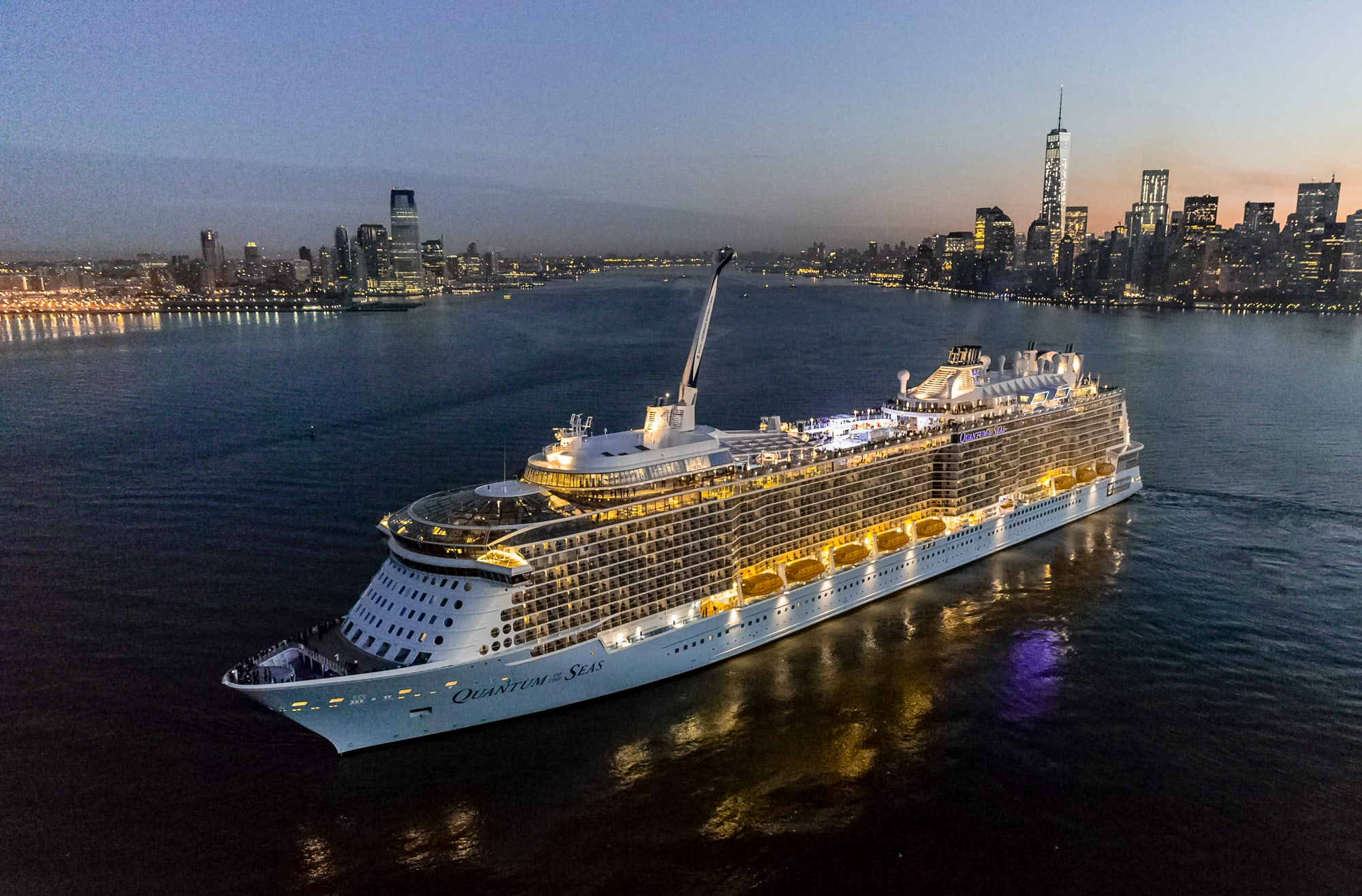 A massive modern cruise ship illuminated against the twilight sky, anchored in New York Harbor