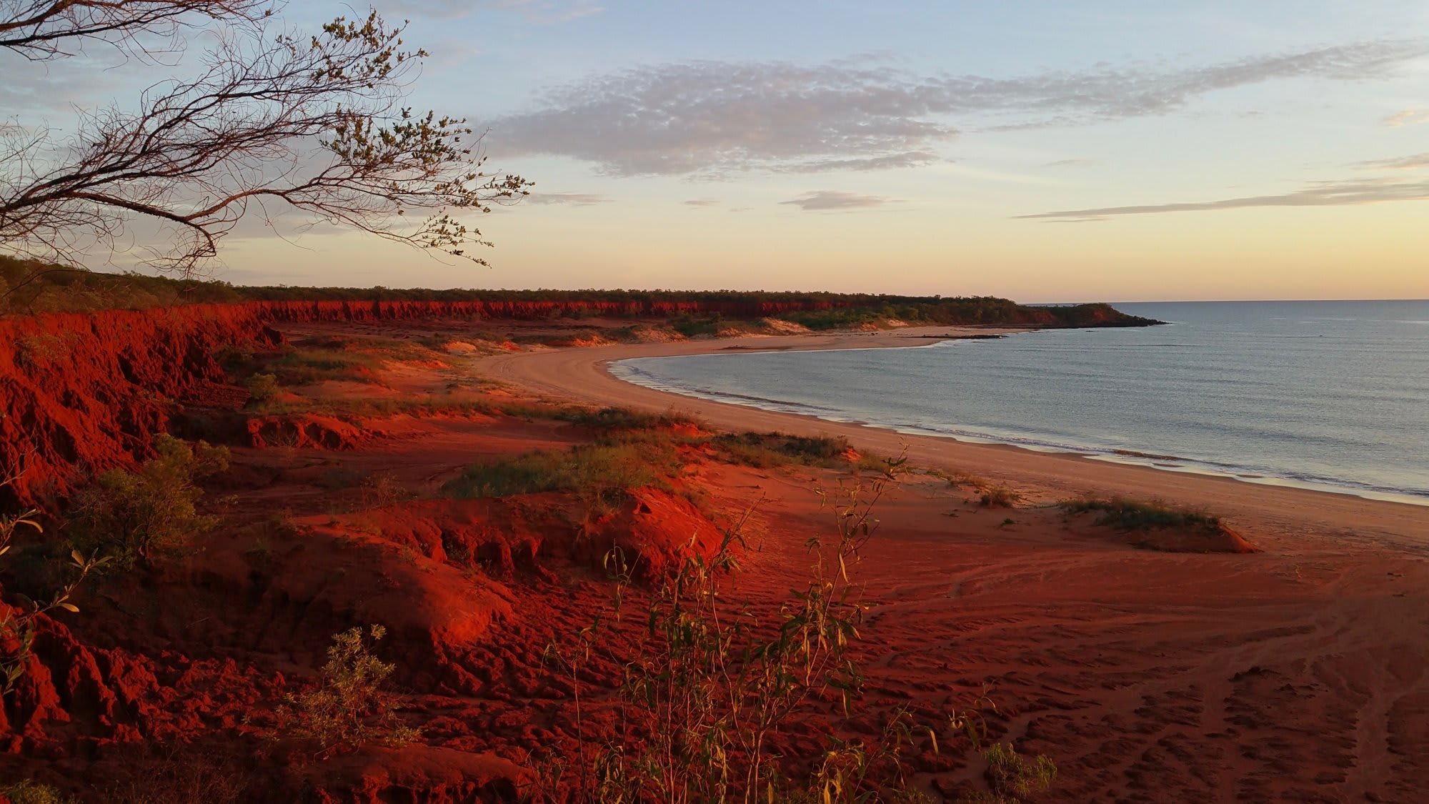 A stunning coastal landscape featuring dramatic red sandstone cliffs overlooking a curved sandy beach and calm waters at dusk. The warm golden light of sunset illuminates the distinctive rust-colored earth formations, creating a striking contrast with the blue-gray ocean and pale sky beyond.