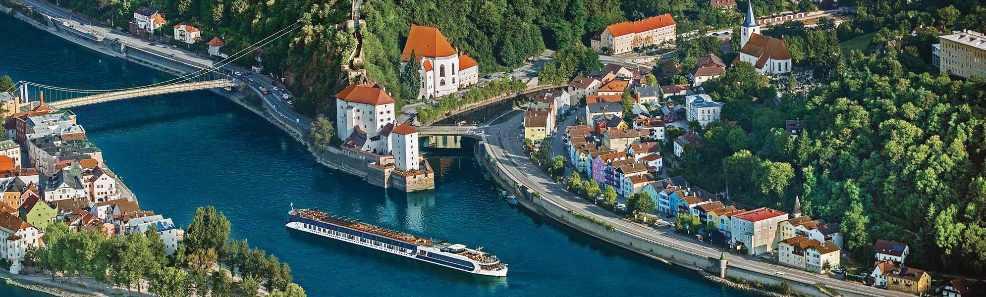 River Cruise on the Danube in Passau An aerial view of a river cruise ship navigating through the picturesque town of Passau, Germany, situated at the confluence of three rivers. The scene showcases the town's distinctive architecture with colorful buildings, baroque churches with orange-tiled roofs, and surrounding forested hillsides, with multiple bridges spanning the blue waters.