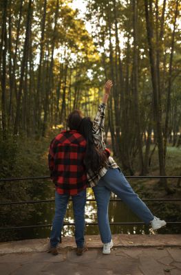 Two people stand side by side near a railing in a wooded area. One person raises an arm and lifts a leg playfully, while the other stands with hands in pockets.