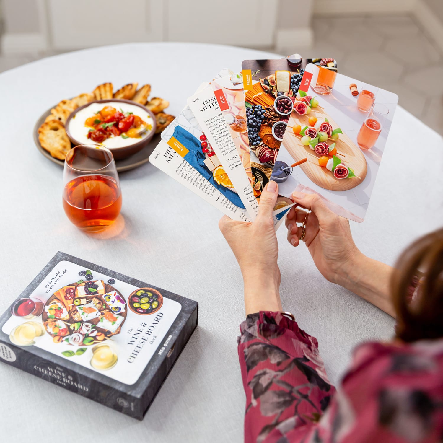 A photo of Meg holding the cards from The Wine and Cheese Board Deck fanned. With a table and wine