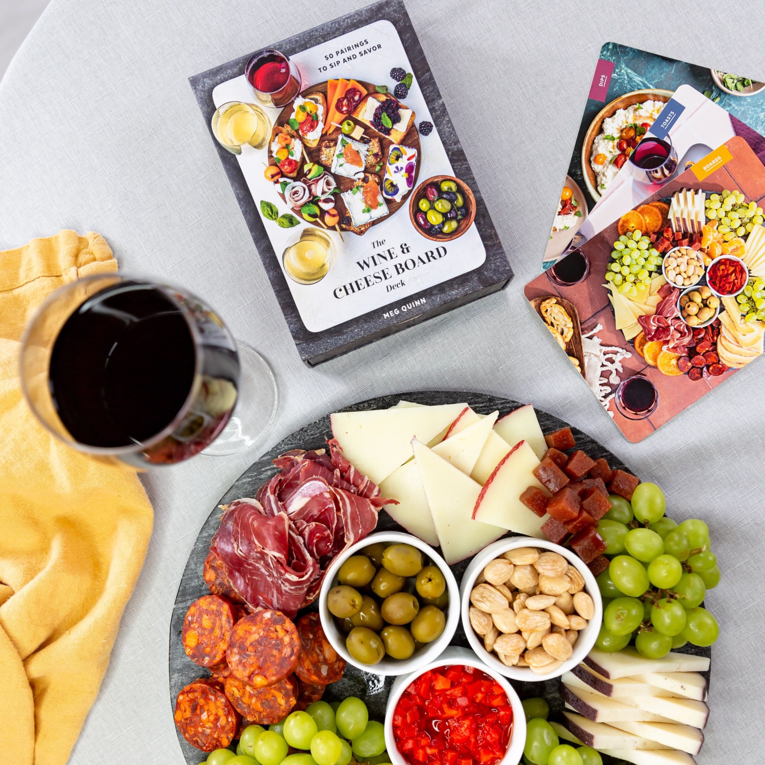 An overhead photo of a cheese board on a table with The Wine and Cheese Board Deck and a glass of wine