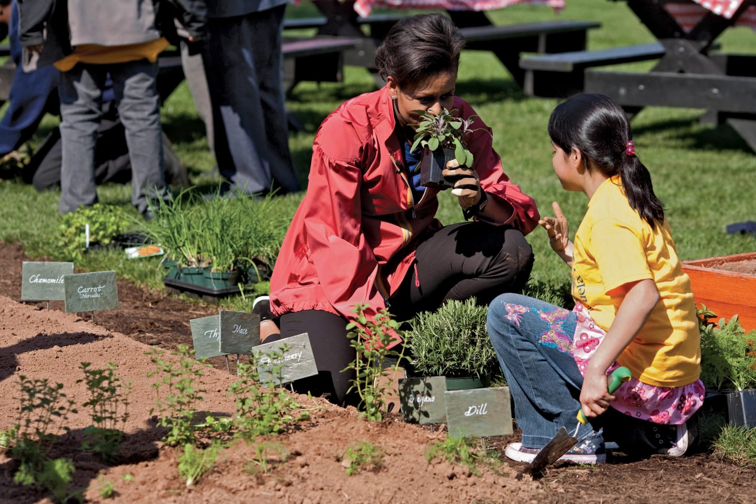 Michelle Obama kneeling in the garden, smelling herbs with a child