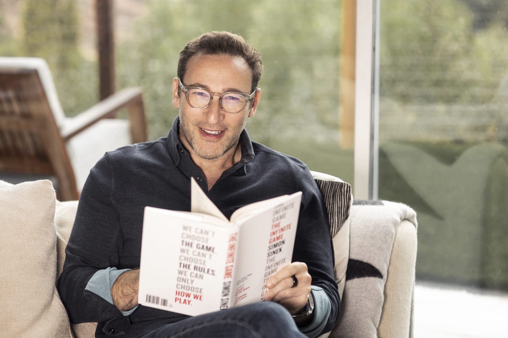 Simon Sinek sitting in a tan chair smiling and holding a copy of The Infinite Game open in his lap. He is wearing a dark blue button down shirt and glasses.