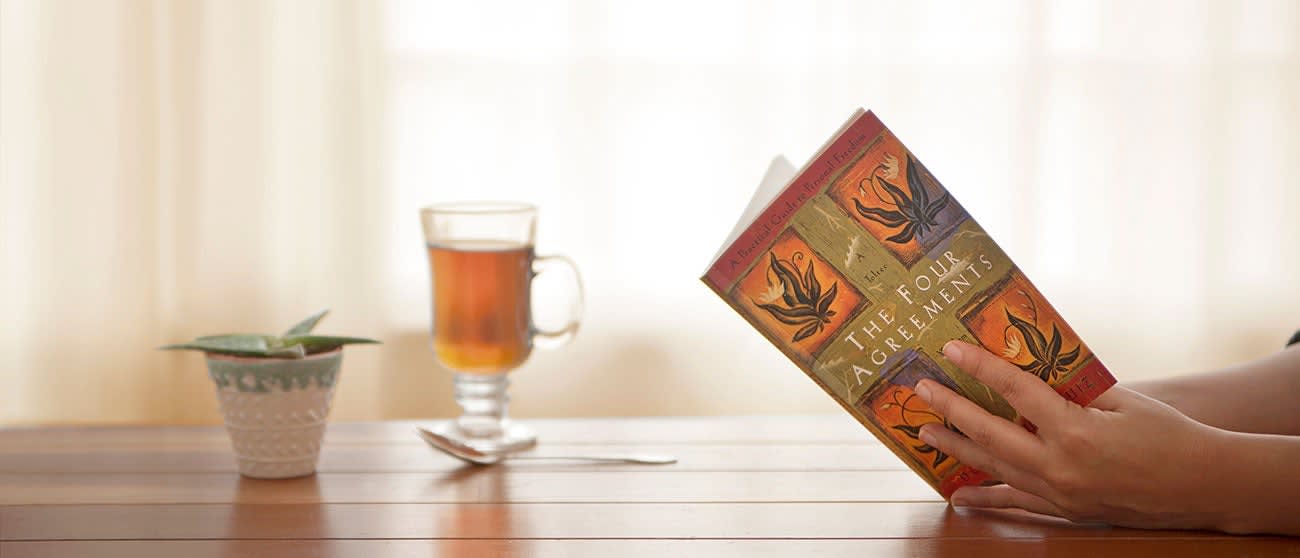 A hand holding the book The Four Agreements with a cup of tea and succulent in the background
