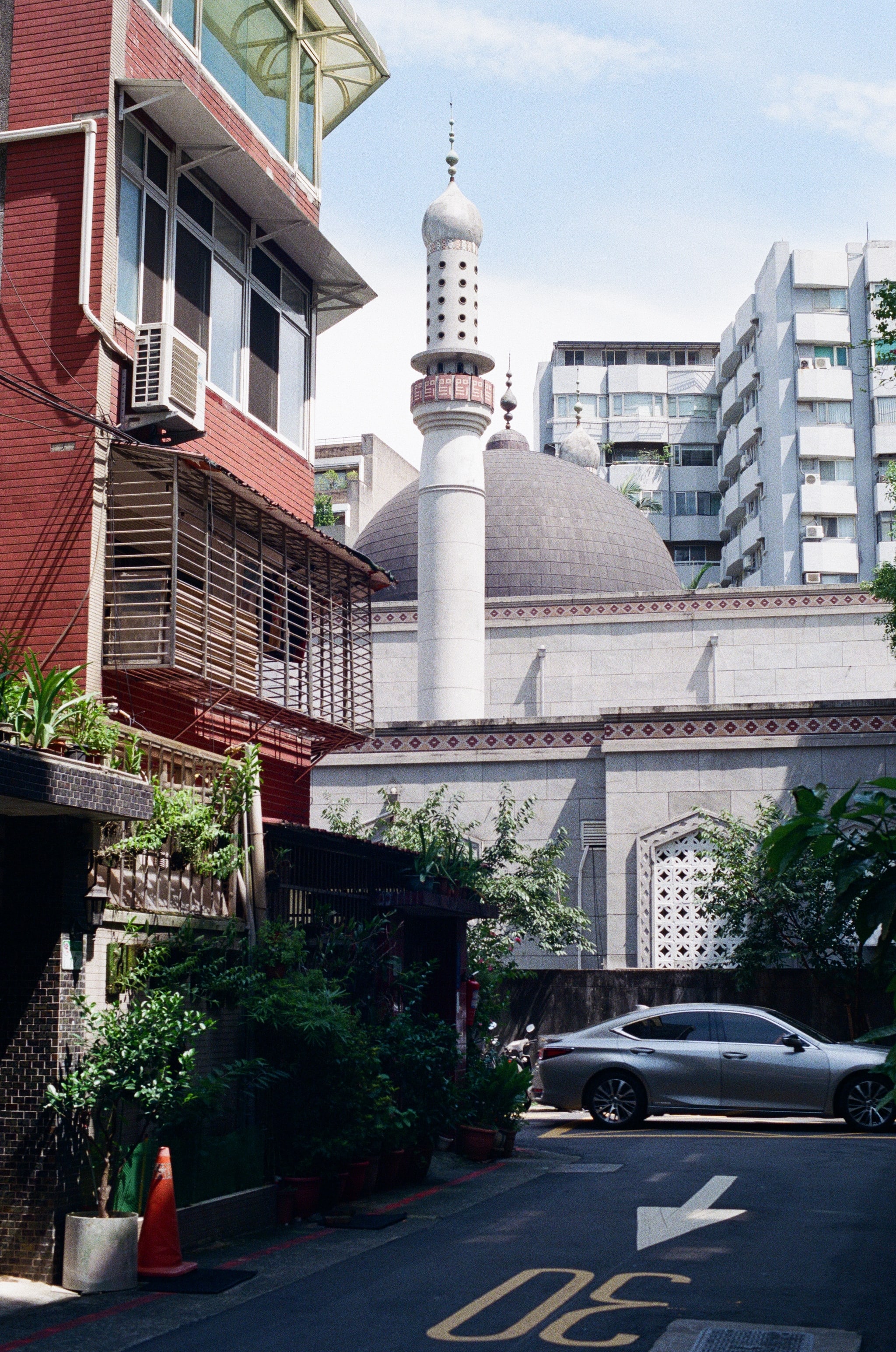 A street scene in a small alley. The frame is dominated by a mosque building in the background. A car drives through the frame in front of the mosque, while a residential apartment building is lit by the midday sun on the left of the shot.