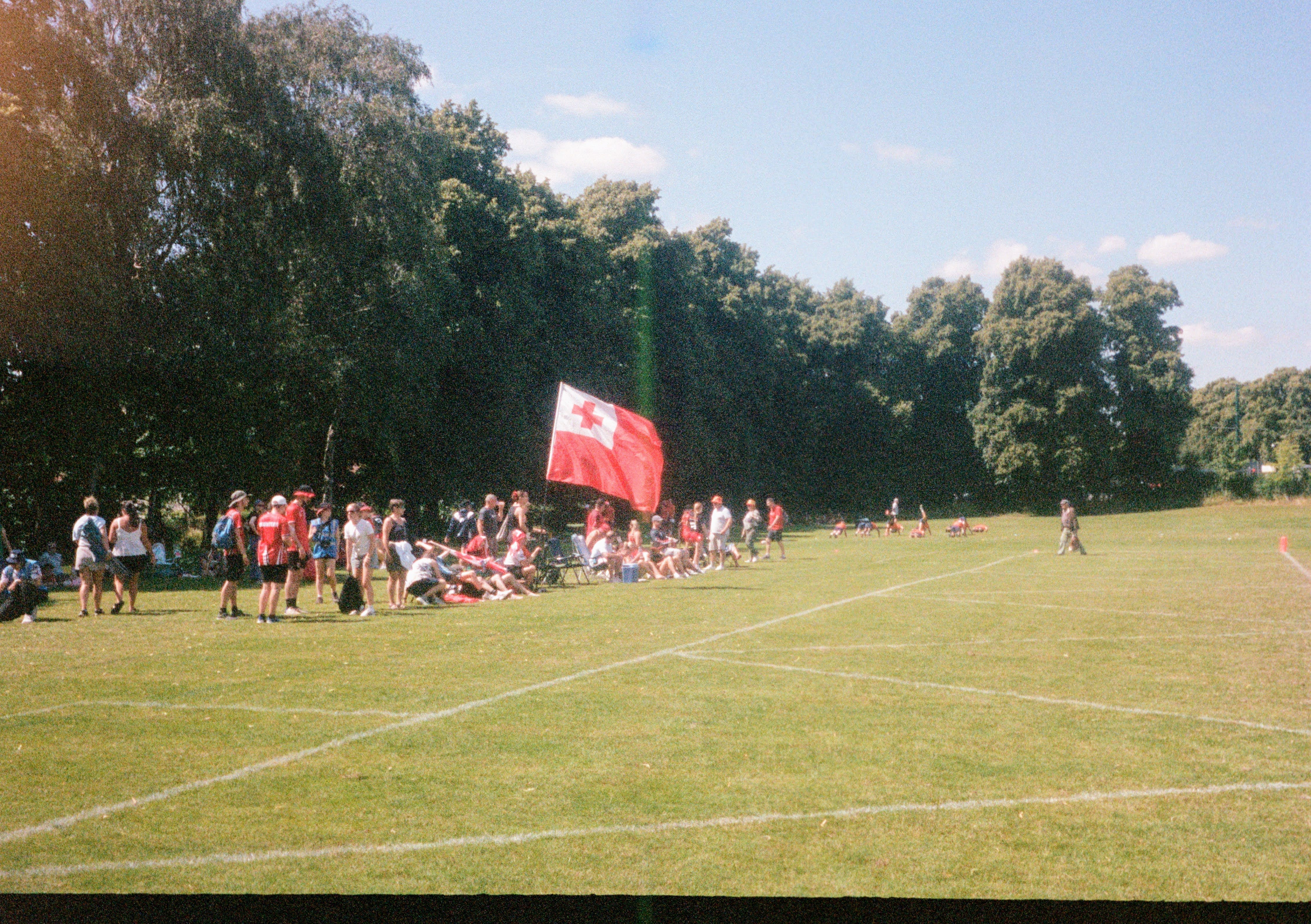 A crowd of people sit by the sideline of a playing field, with a large Tongan flag in the center of their group.