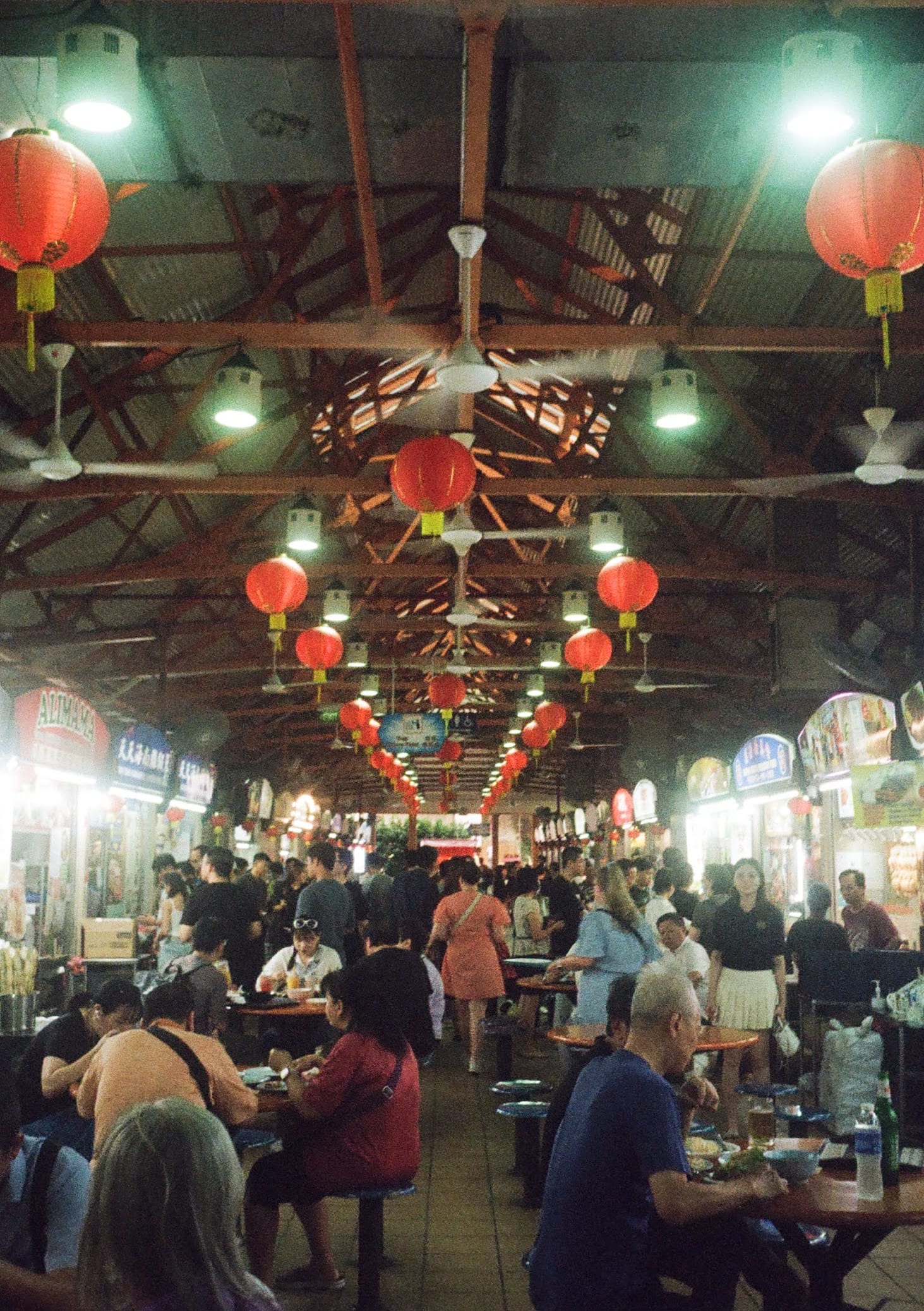 People sit at round tables eating, while others walk between carrying meals. The roof above is lined with red lanterns.