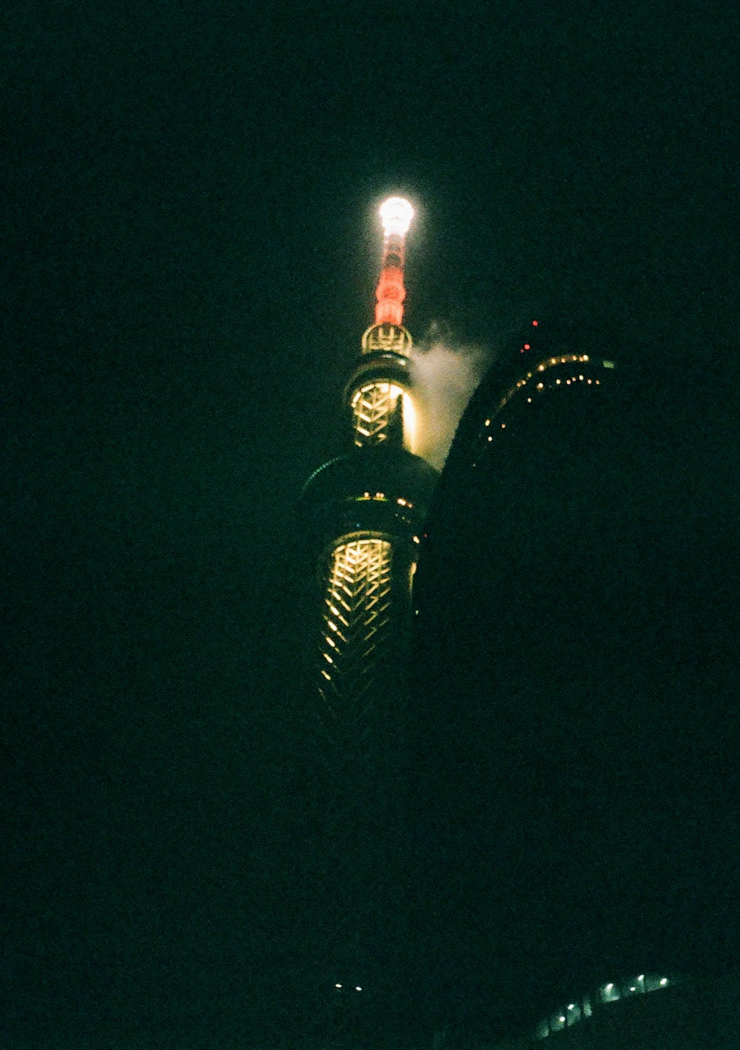 The spire of a building is lit up with clouds floating around it. The building is partially blocked by a low building in the foreground.