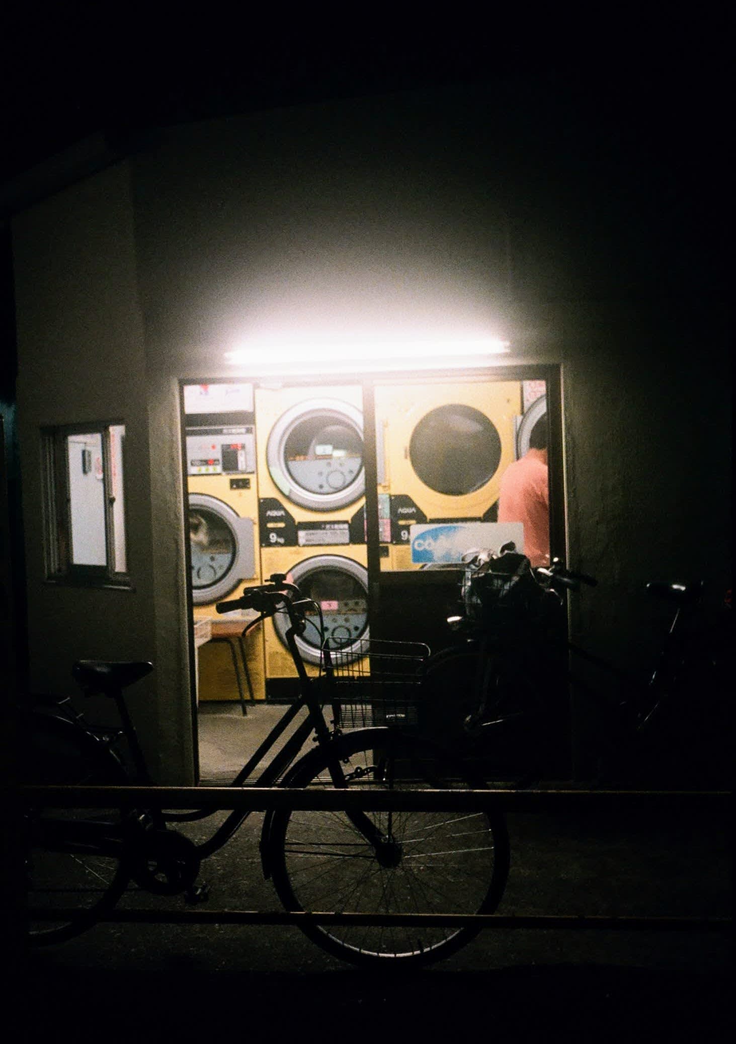 A bike sits at the entrance of a laundromat, the machines behind it brightly lit against the night.