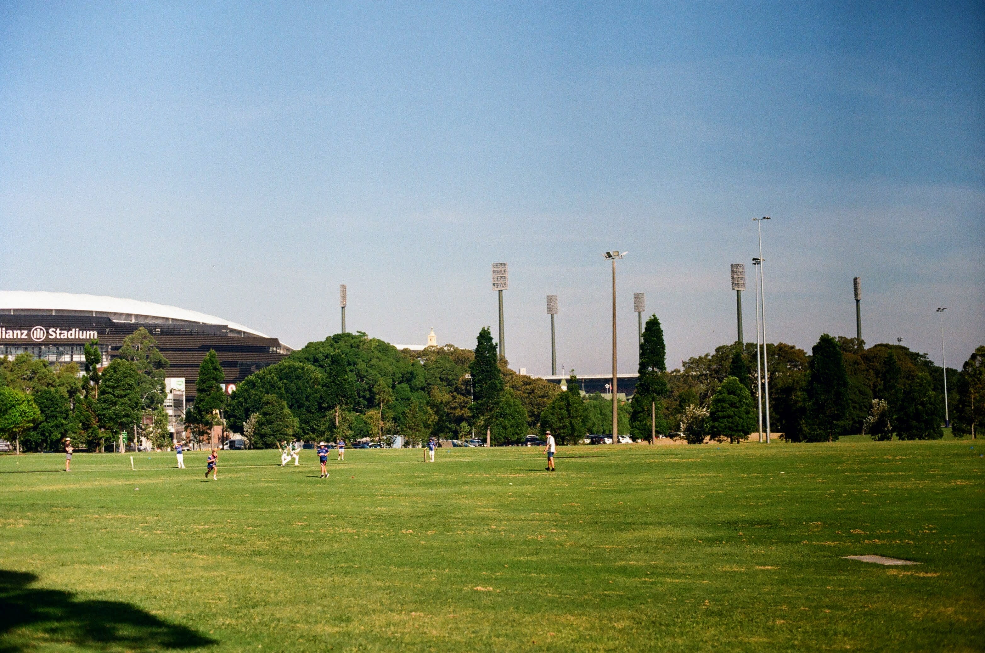 Kids playing cricket in a large open field, with blue skies above. Two large stadiums dominate the background of the shot.