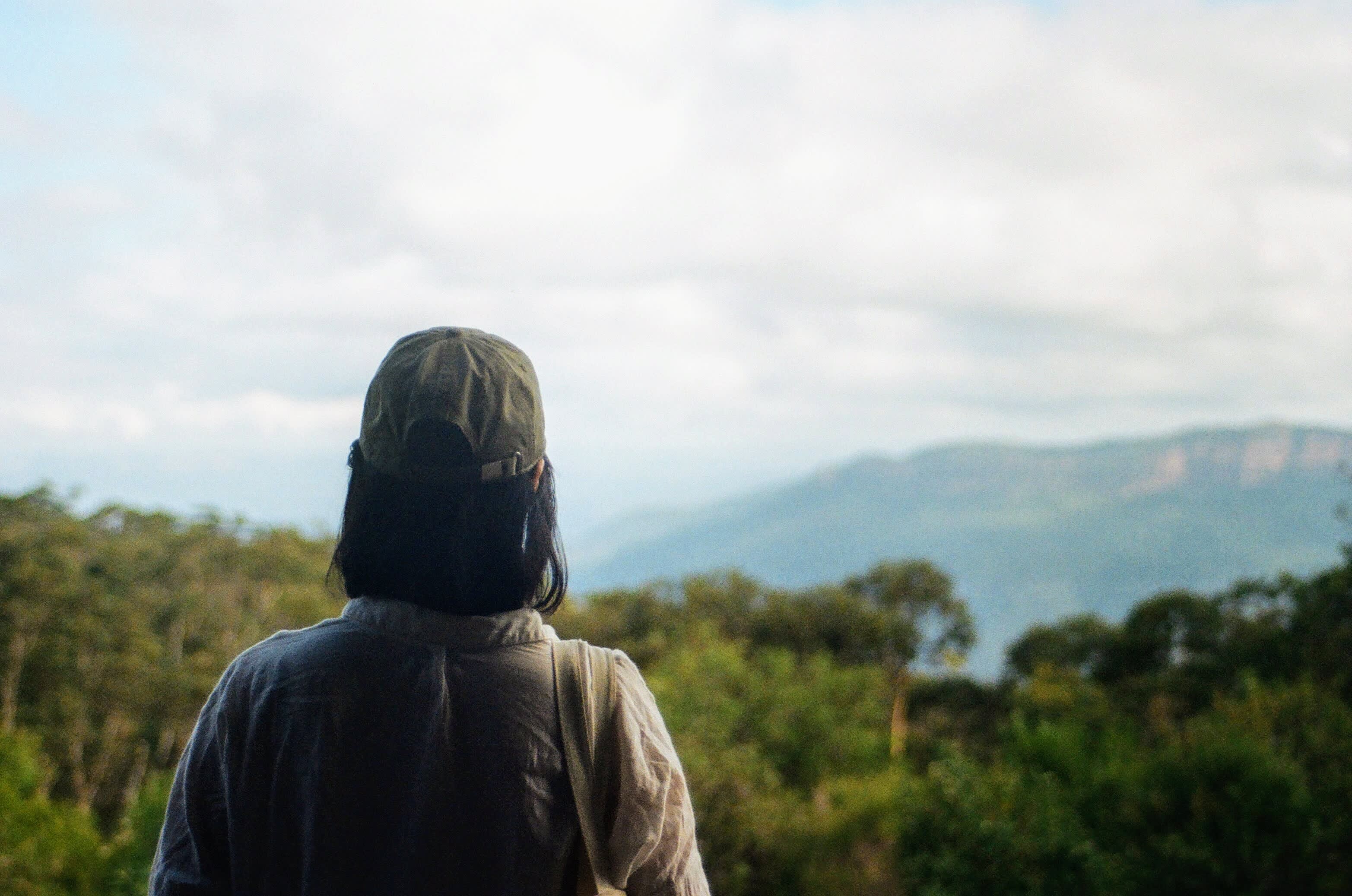 A female wearing hiking clothes has her back to the camera. She is looking out over green trees that extend out over hills in the background.