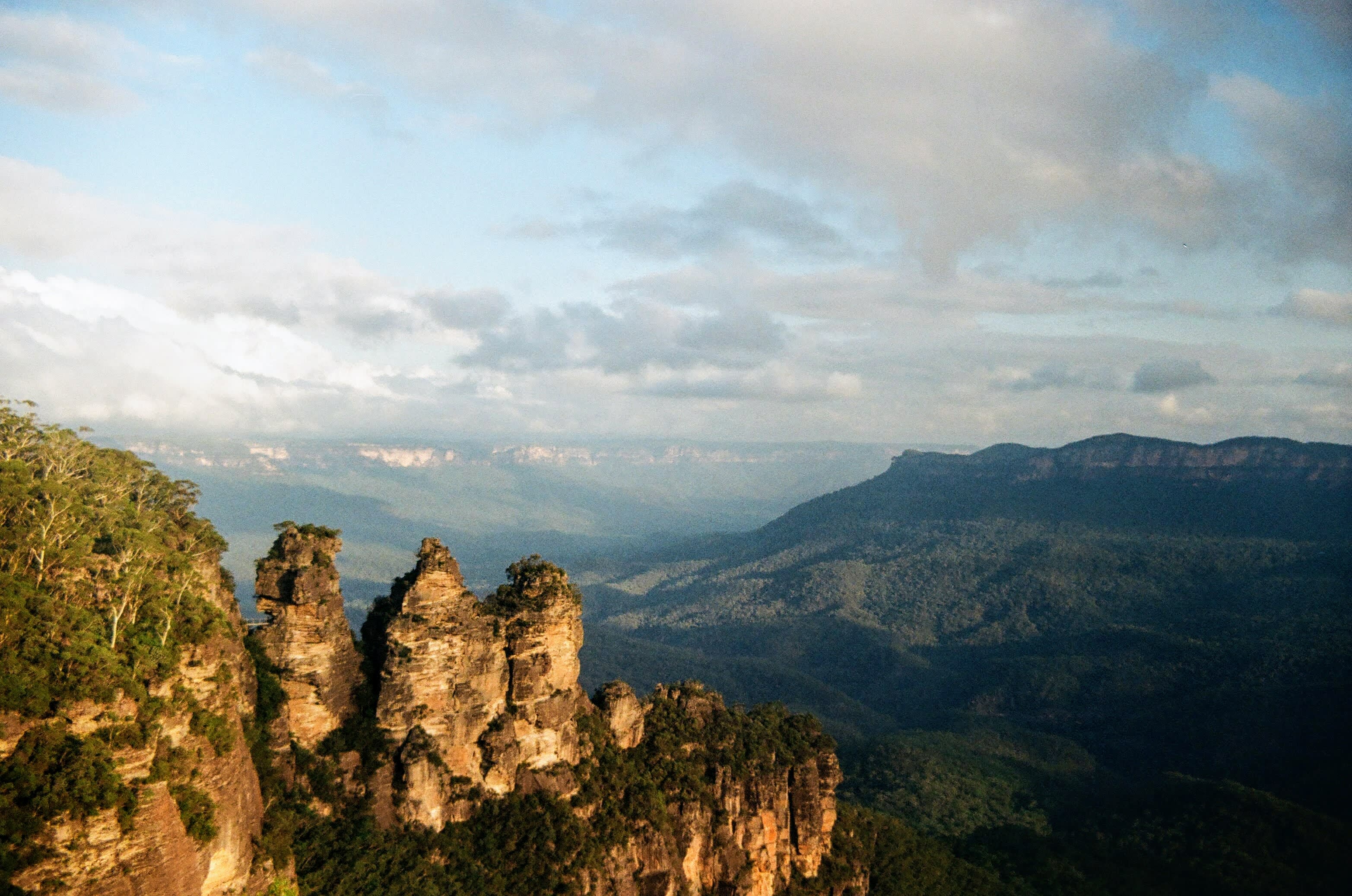 The Three Sisters rock formation stands in the foreground, illuminated by soft light of the setting sun against a backdrop of a vast, hazy mountain range under a partly cloudy sky.