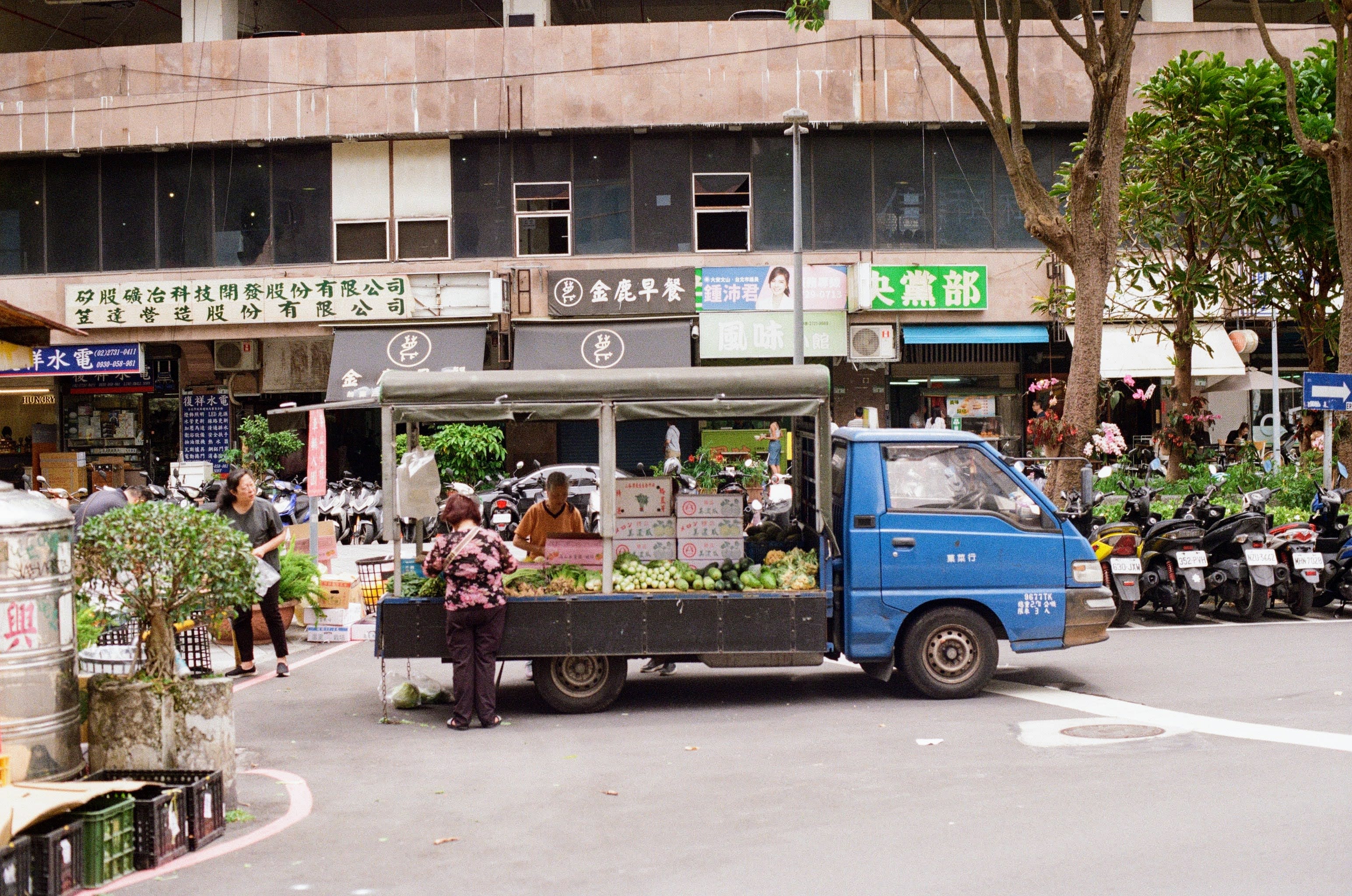 A blue truck is in the center of the picture with its back open with fruits and vegetables on display. An eldery lady is looking at the produce on display.