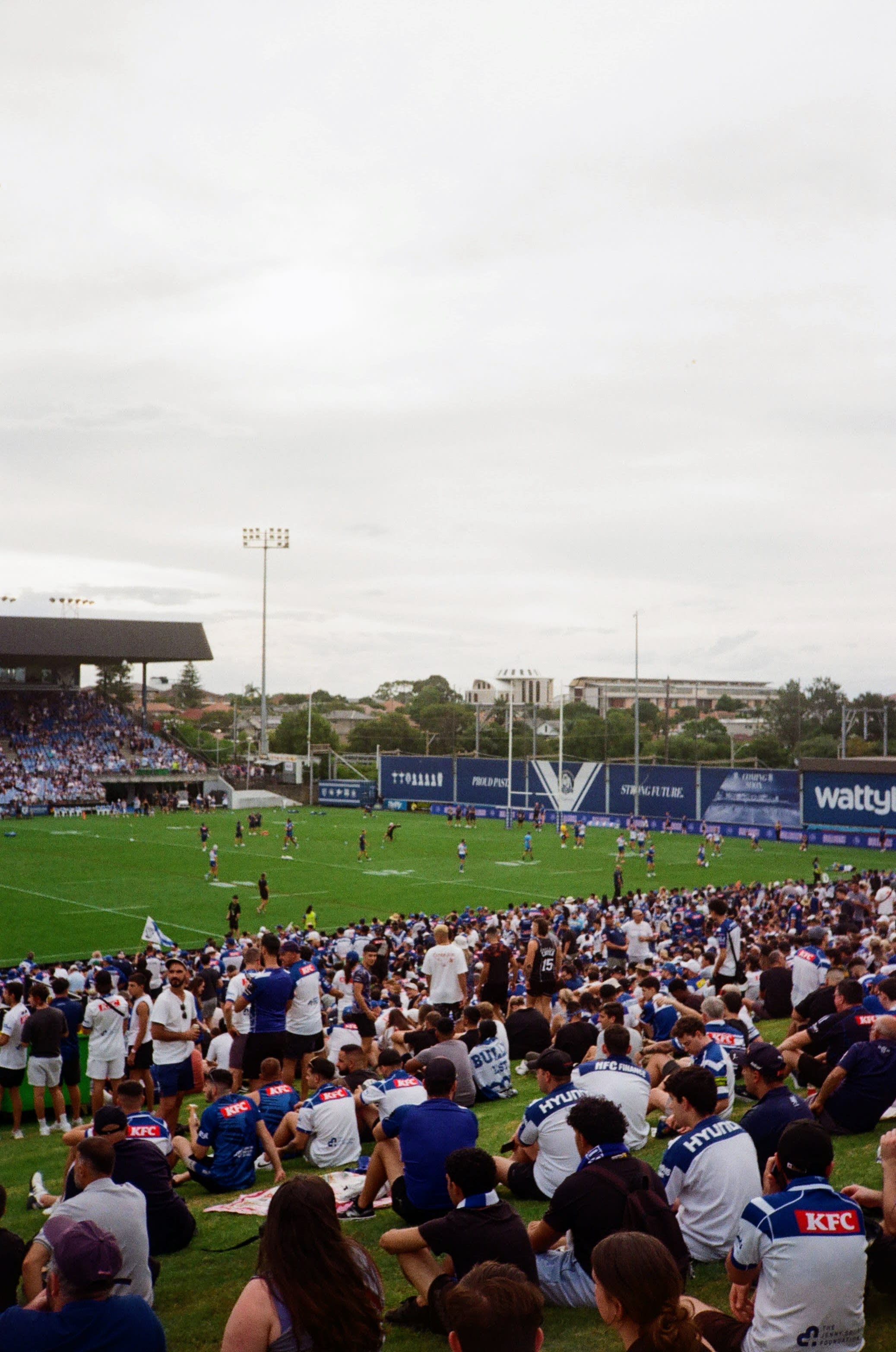 A crowd of spectators wearing blue and white sit on a hill watching a game of Rugby League.