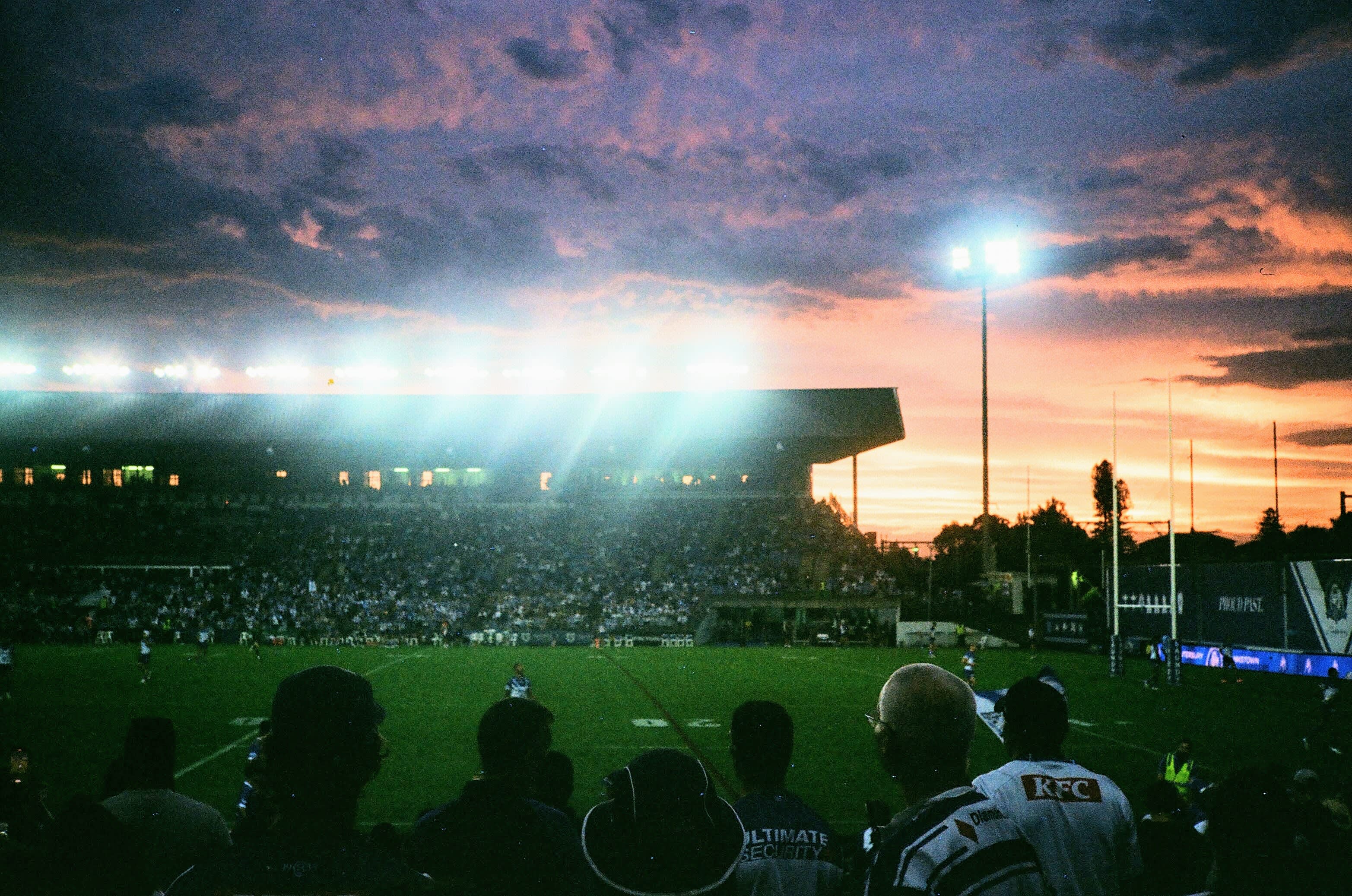 Overlooking a Rugby field, towards a grandstand that is casting light onto the field. The sky is a mix of orange and purple hues as the sun sets in the background.