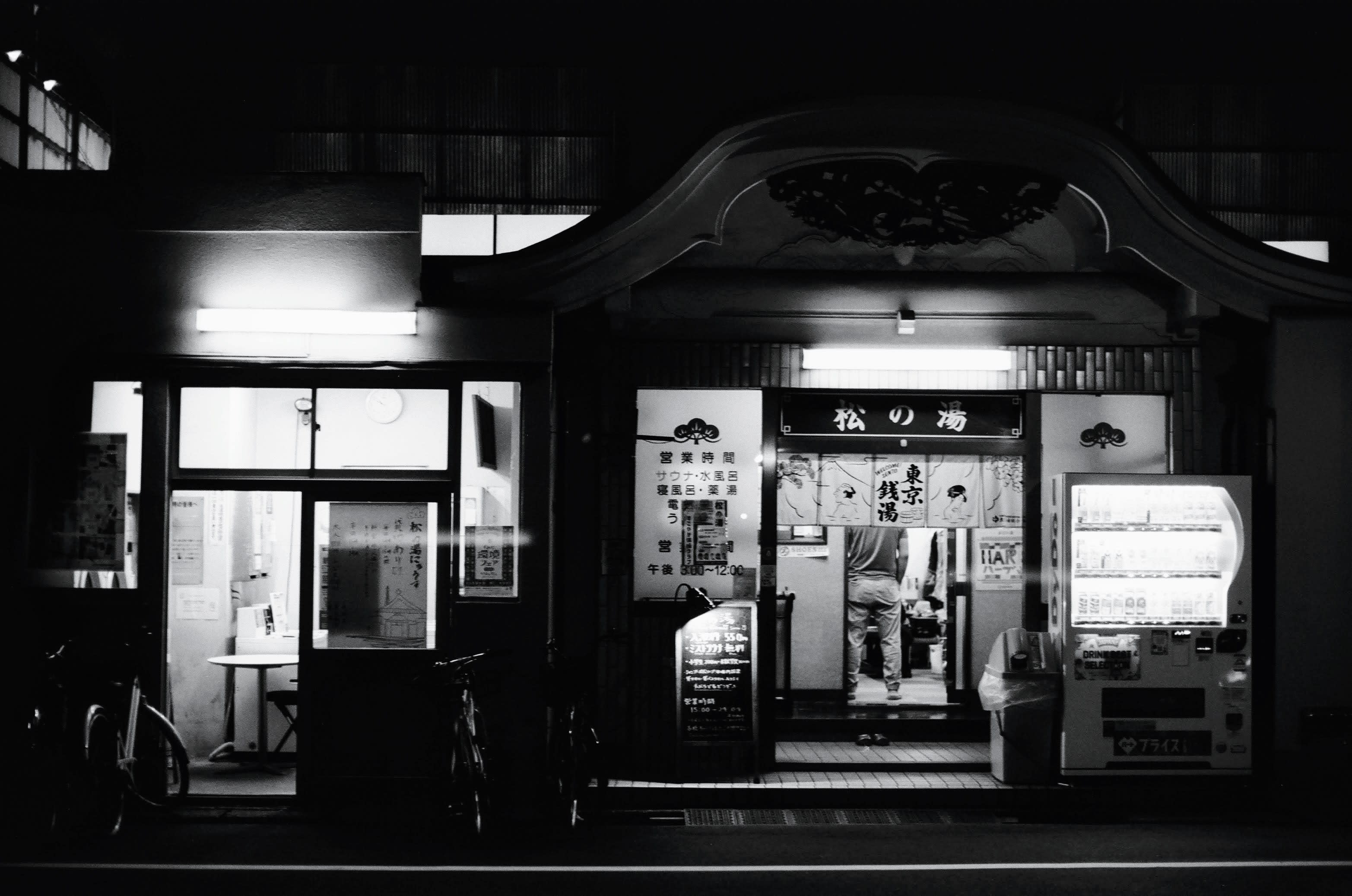 The entrance to a Japanese bath house, with a vending machine on the right and laundry room on the left illuminating the black and white image.