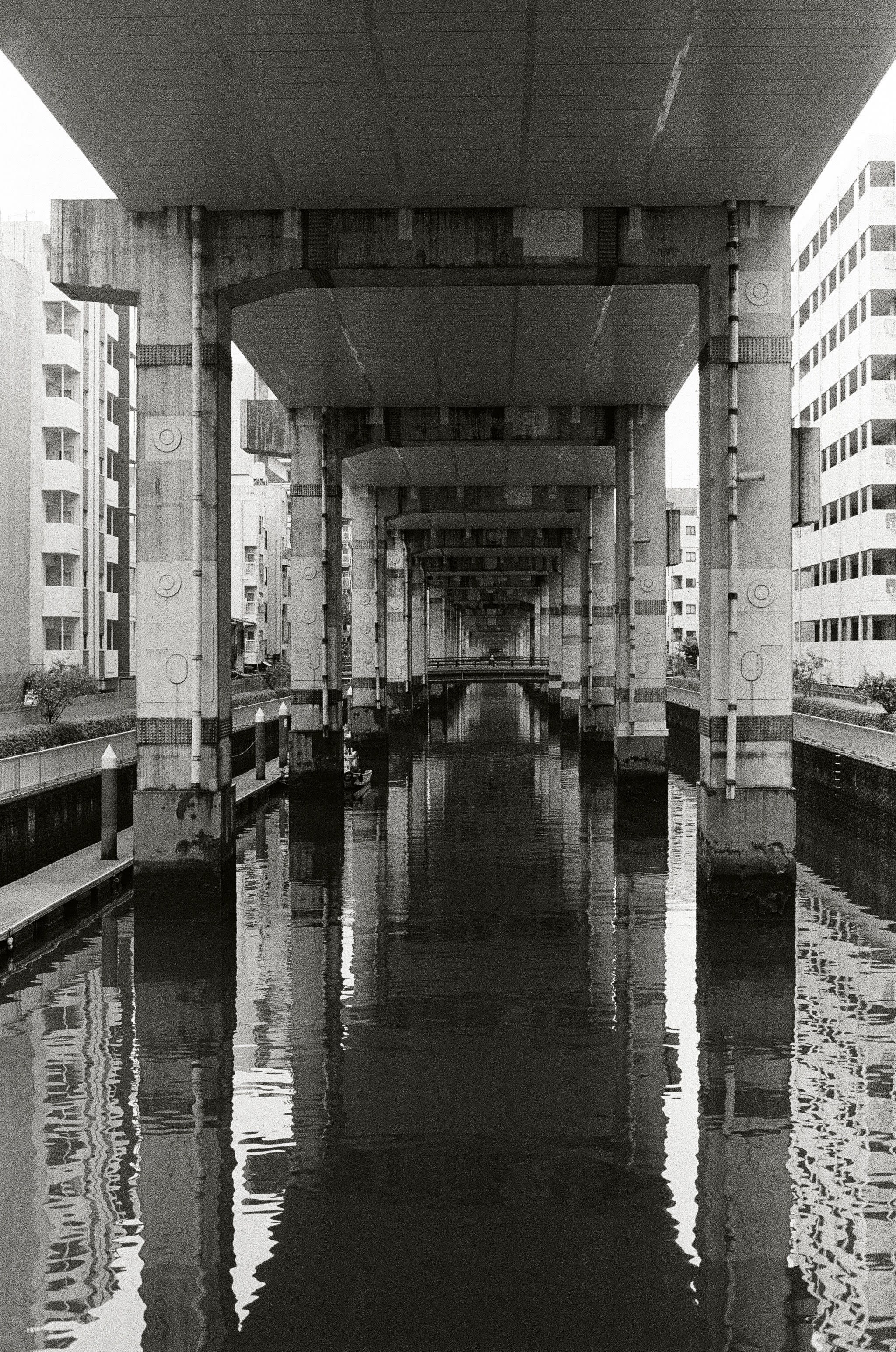 Supporting pylons for an overhead expressway continue in a sequence into the distance. They are reflected in a canal that runs below the road.