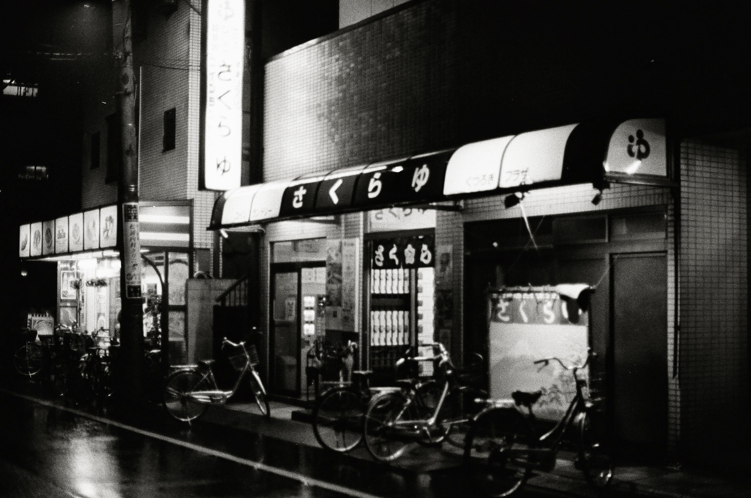 Bicycles are parked outside a brightly lit Japanese bath house. A sign with the name of the venue is above the door. A laundry room is lit up on the left of the entrance.