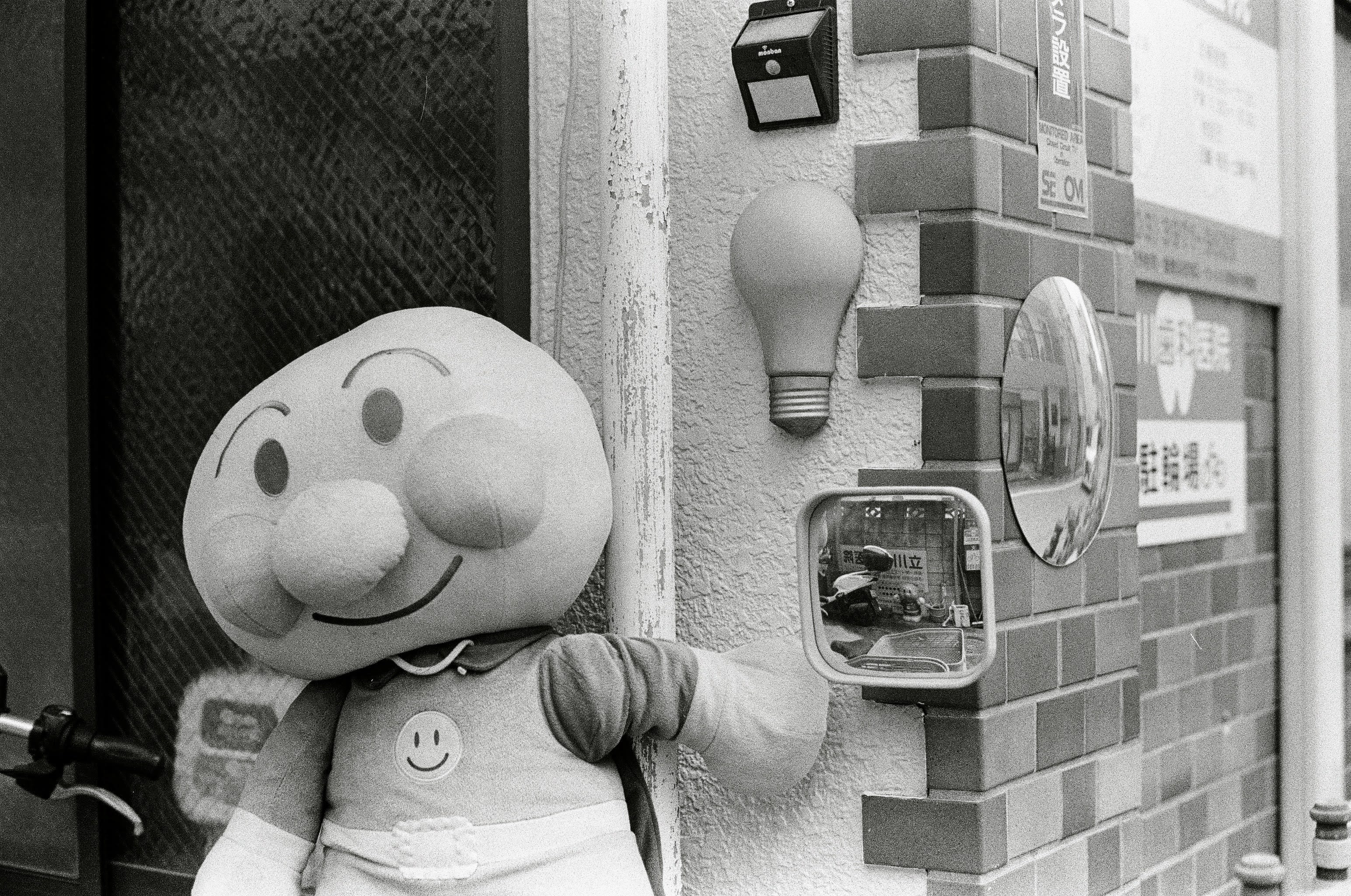 A large Anpanman doll sits outside a house in Tokyo. Next to it is a small mirror which has another smaller Anpanman character reflected in it.