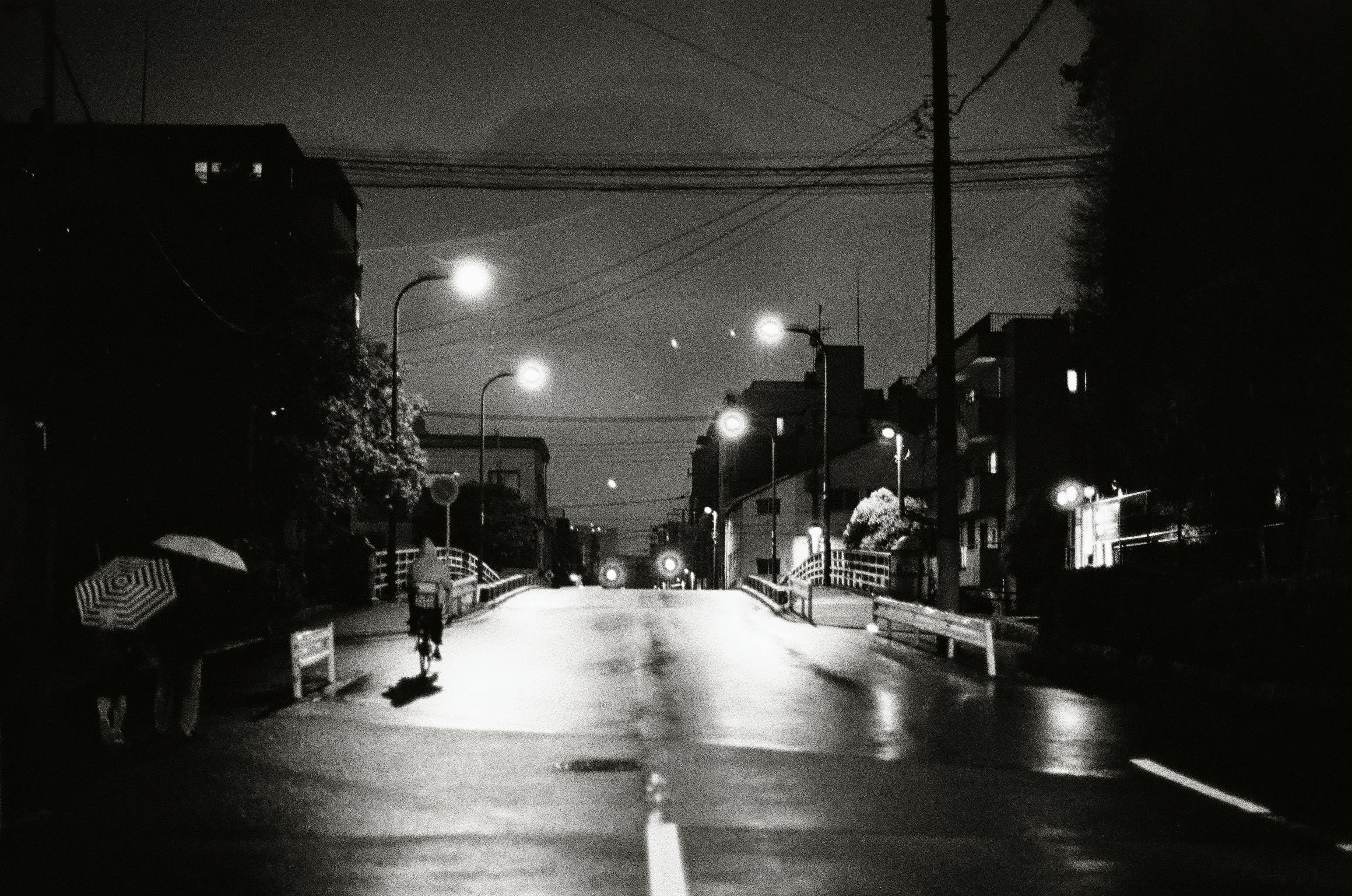 A person in a rain jacket rides a bike over a bridge, while a lady and child walk behind them under and umbrella. The road glistens as the street lights reflect off the wet surface.