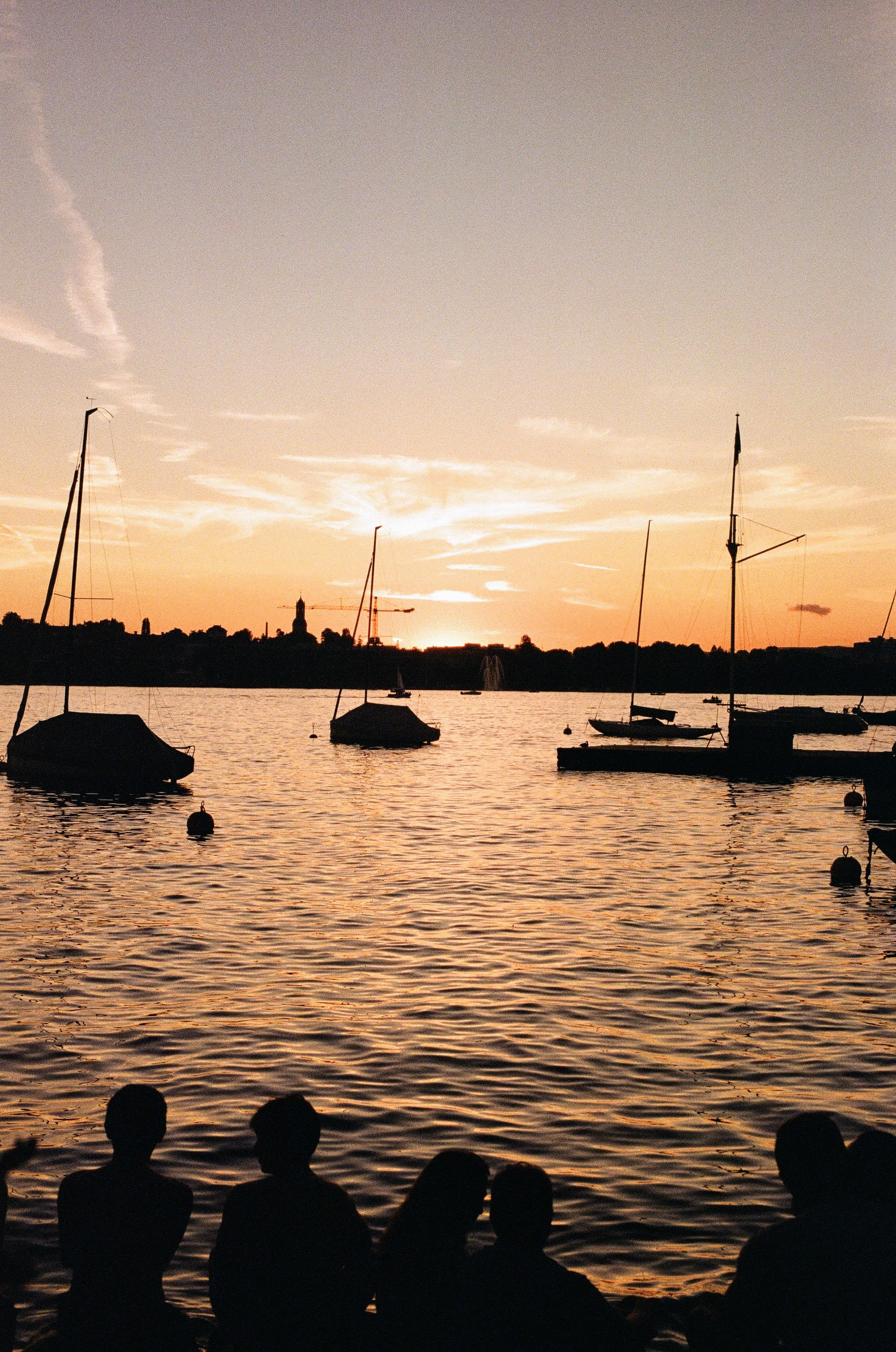 Photo of an orange sky and orange lit lake at sunset. Silhouettes of people sitting on the lake shore are in the foreground, with silhouetted boats on the lake, and a city skyline in the distance.
