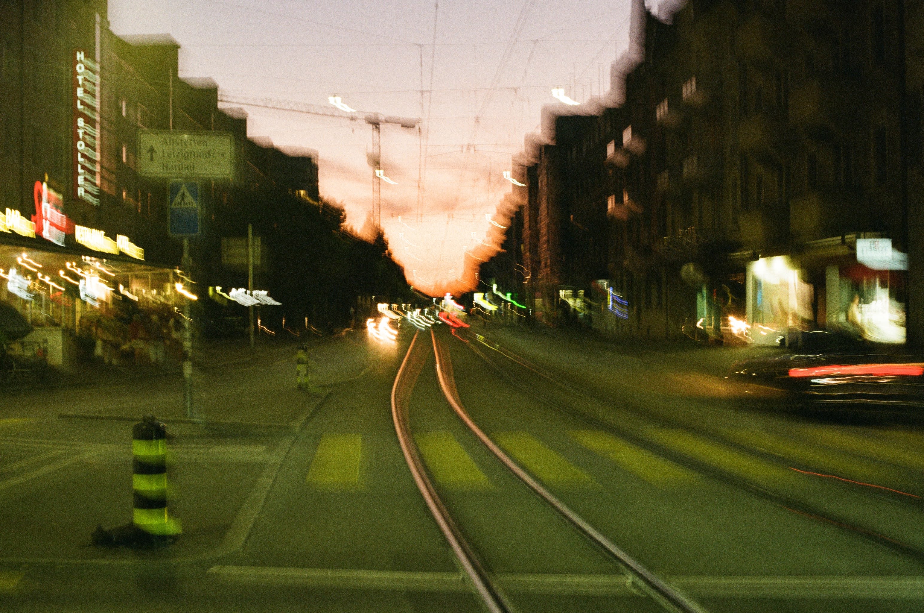 A blurry image looking down a road towards a sunset sky. Car lights and street lights are blurred lines across the shot.