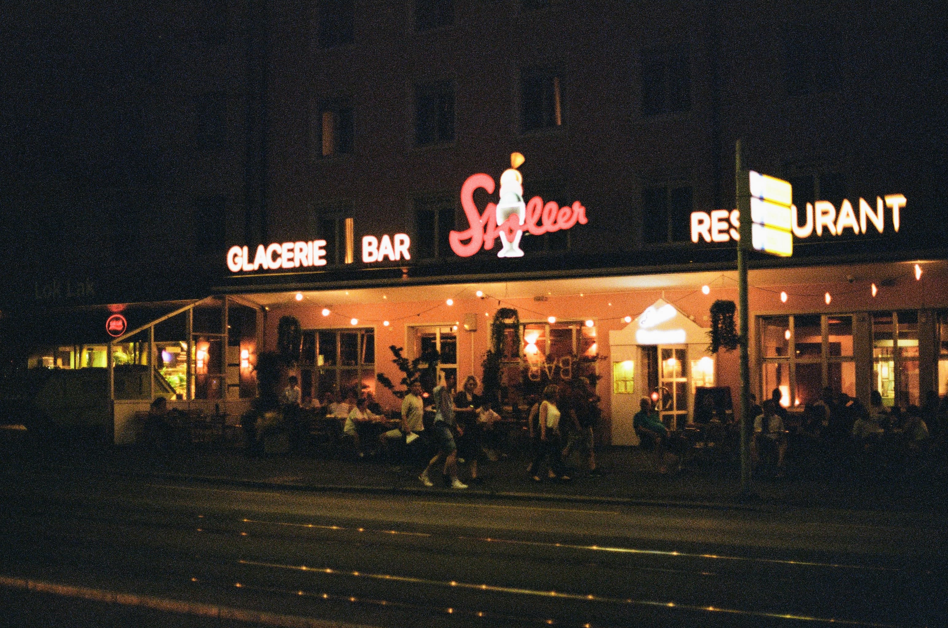 A night shot looking across the road at a bar and ice cream palor with people walking past patron who are sitting outside.