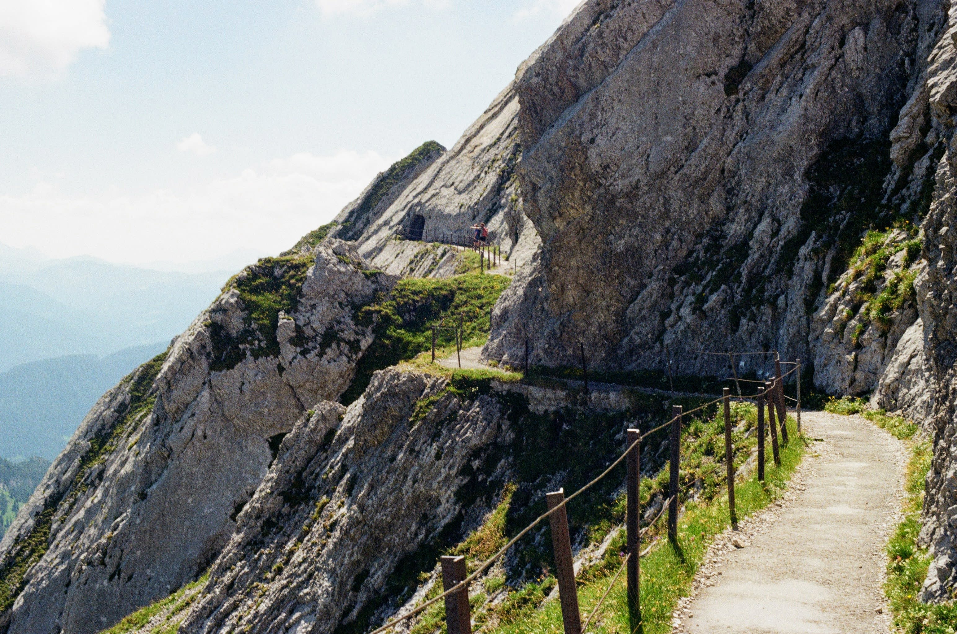 A hiking trail cuts through the rock on the side of a mountain. Two hikers are in the far distance.