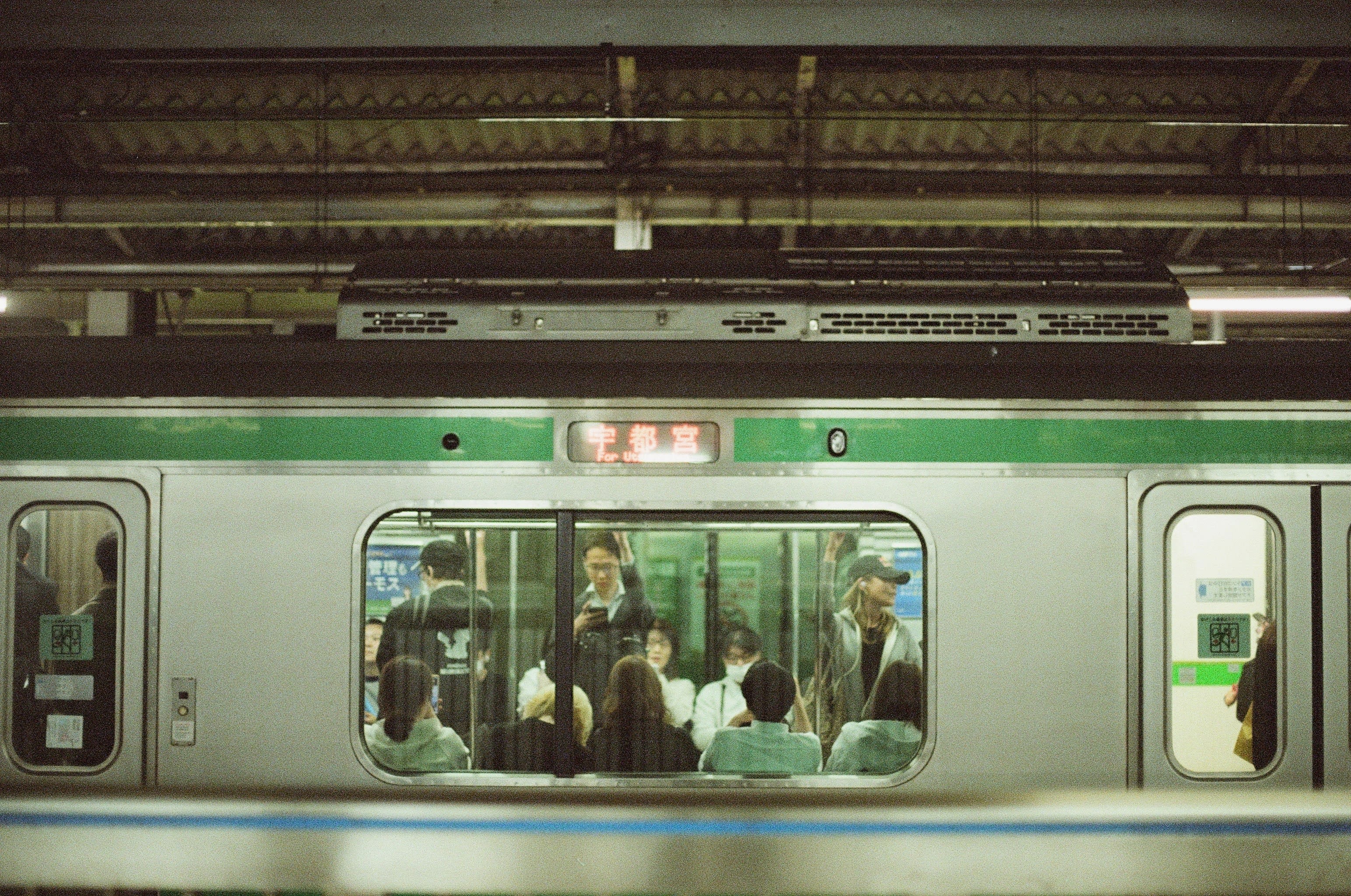 The photograph looks through the window of a passenger train. The backs of seated passengers are seen in the foreground, while standing passengers are centered in the shot. One man looks at his phone, while a women looks away from the camera.