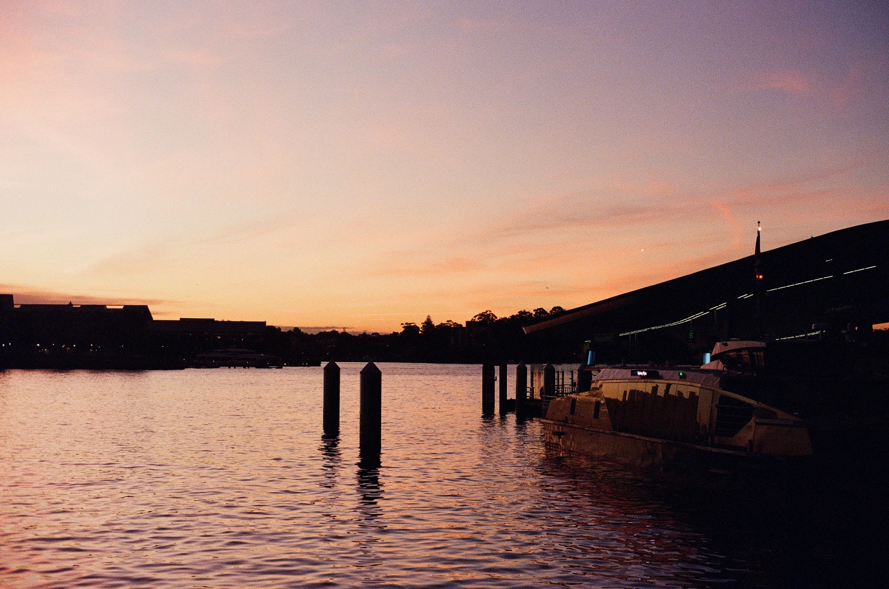 A ferry is docked on the right of the frame, in the shade. The shot is dominated by a view over a body of water, illuminated by the orange glow of the setting sun.