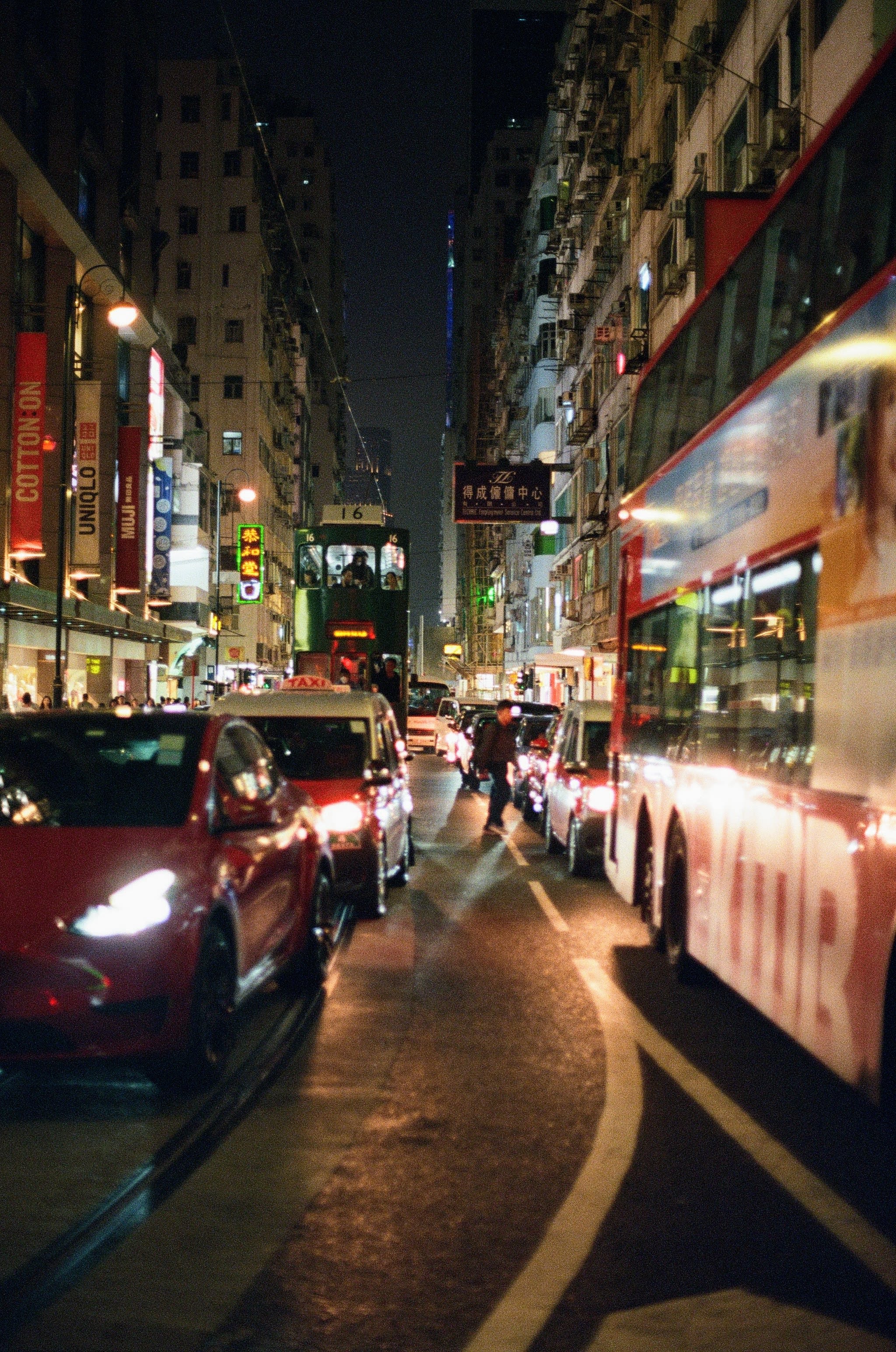 Looking down a busy street in Hong Kong. A red double-deck bus dominates the right of the frame, while the left is filled with two red cars and an approaching tram. The scene is shot at night.