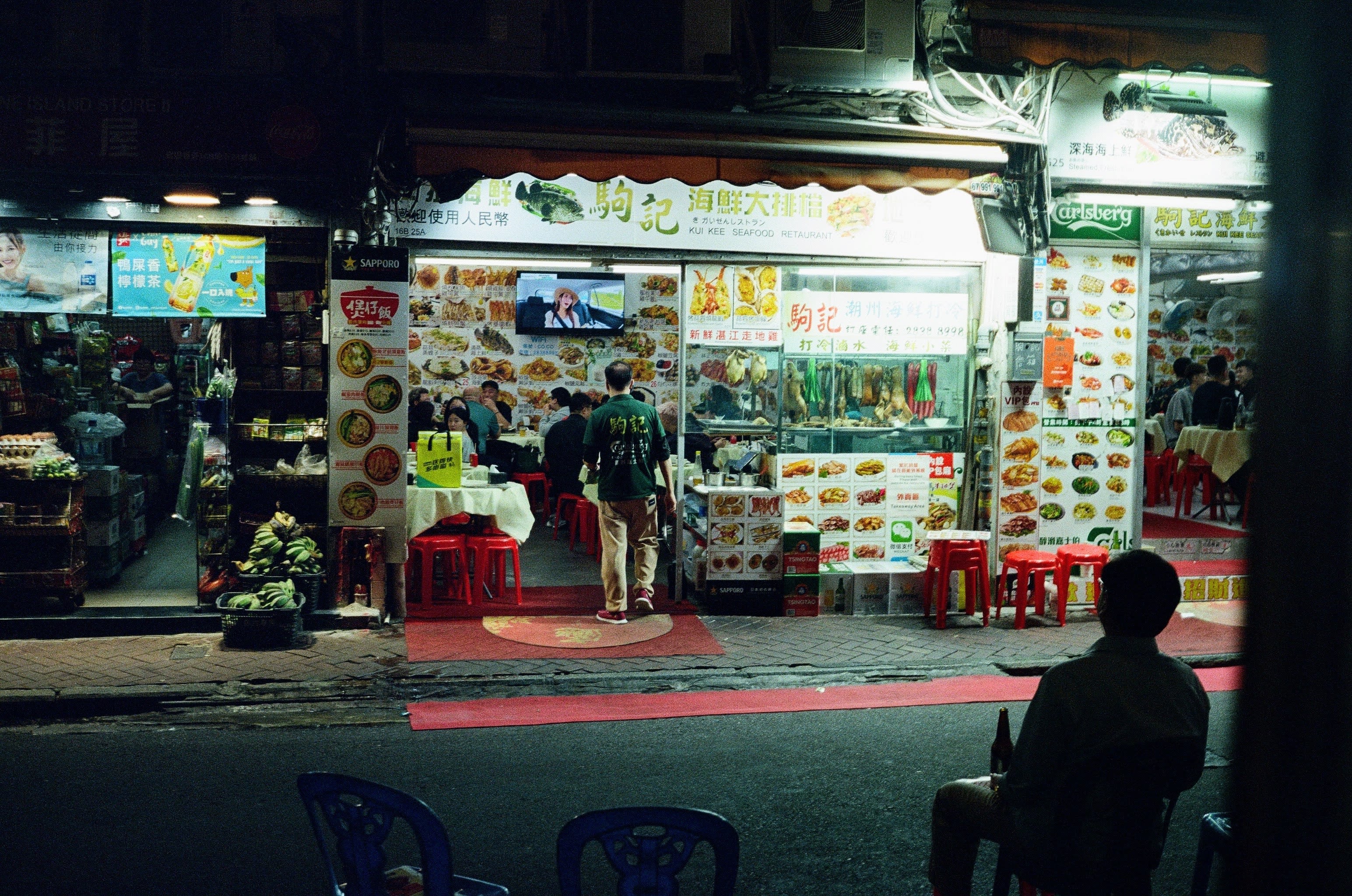 A man sits in the foreground of the scene to the right of the photographer. It is night, and the man is seated in the dark, his back to the camera, beer in hand looking towards a brightly lit restaurant and TV.
