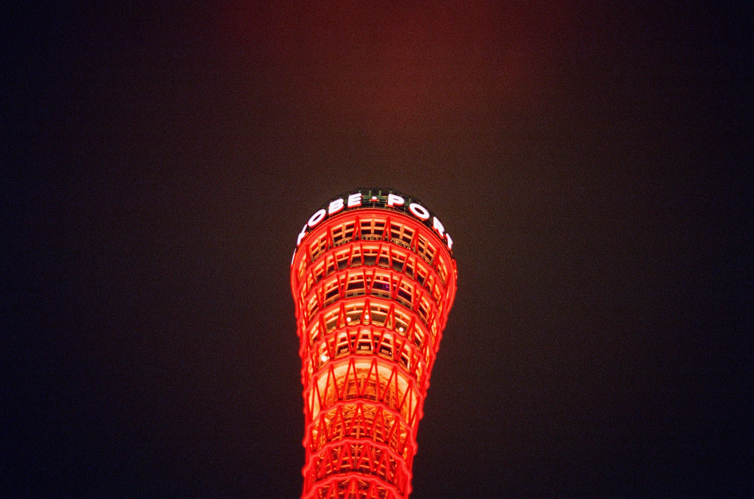Looking up towards a red tower illuminating the night sky. White wording on the top of the tower reads Kobe Port.