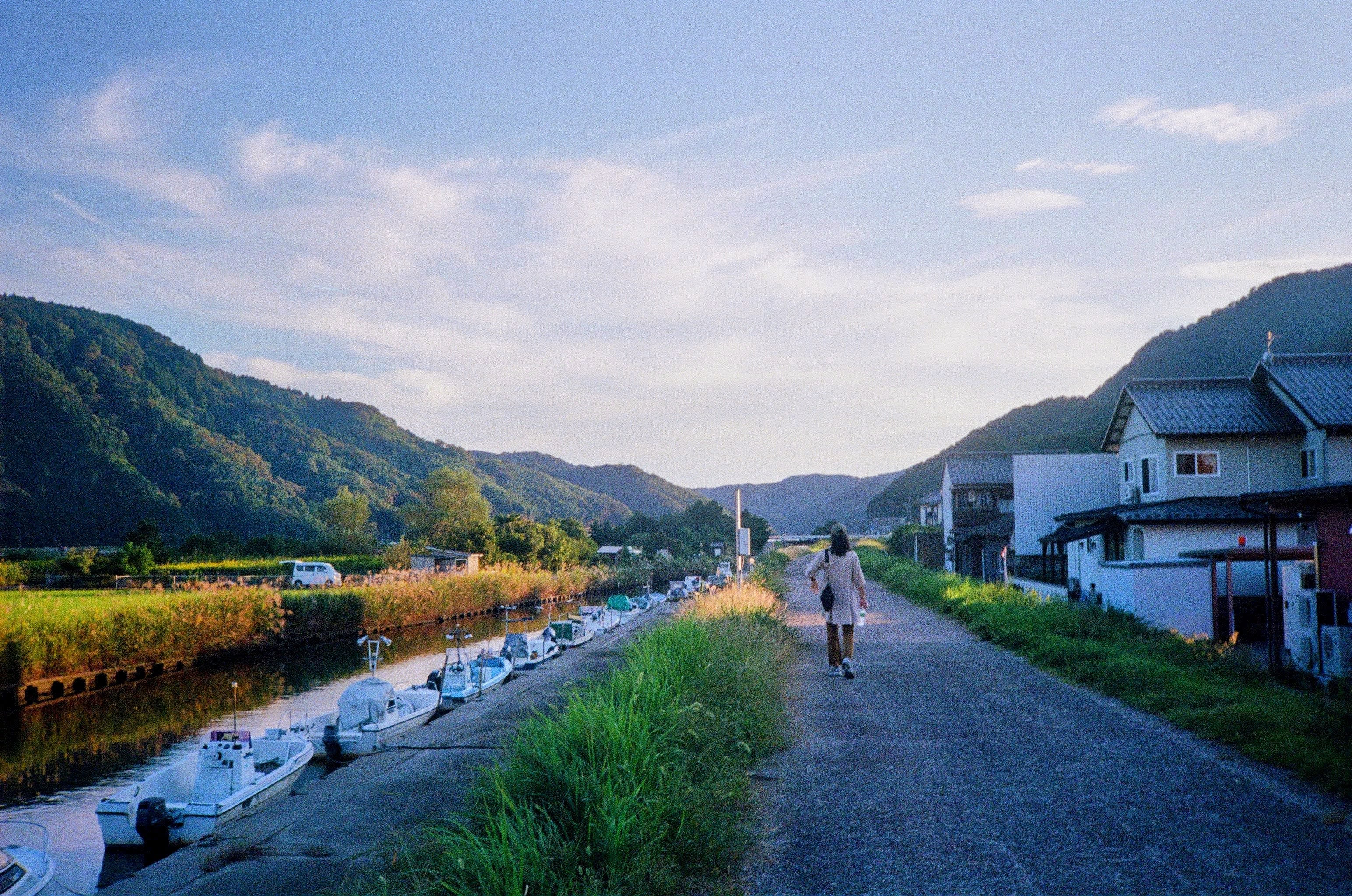 A woman walks away from the camera on a path next to a stream. Houses are on her right, while across the stream is a field that is brightly lit in the afternoon sun. Mountains frame both sides of the shot.