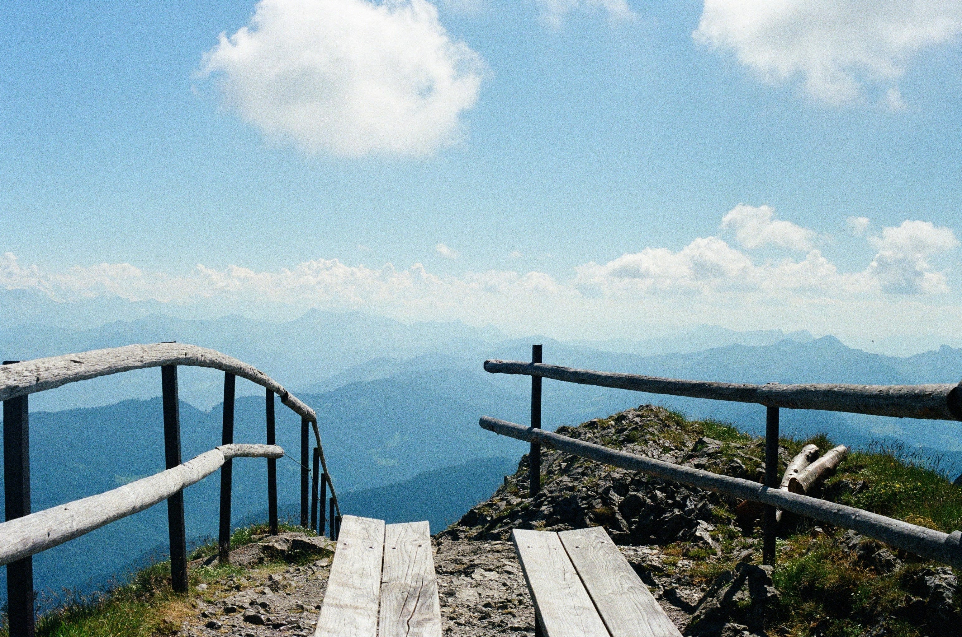 View seated on a bench, looking through a narrow opening between wood railings towards mountains in the distance. The path leading from the benches falls off sharply.
