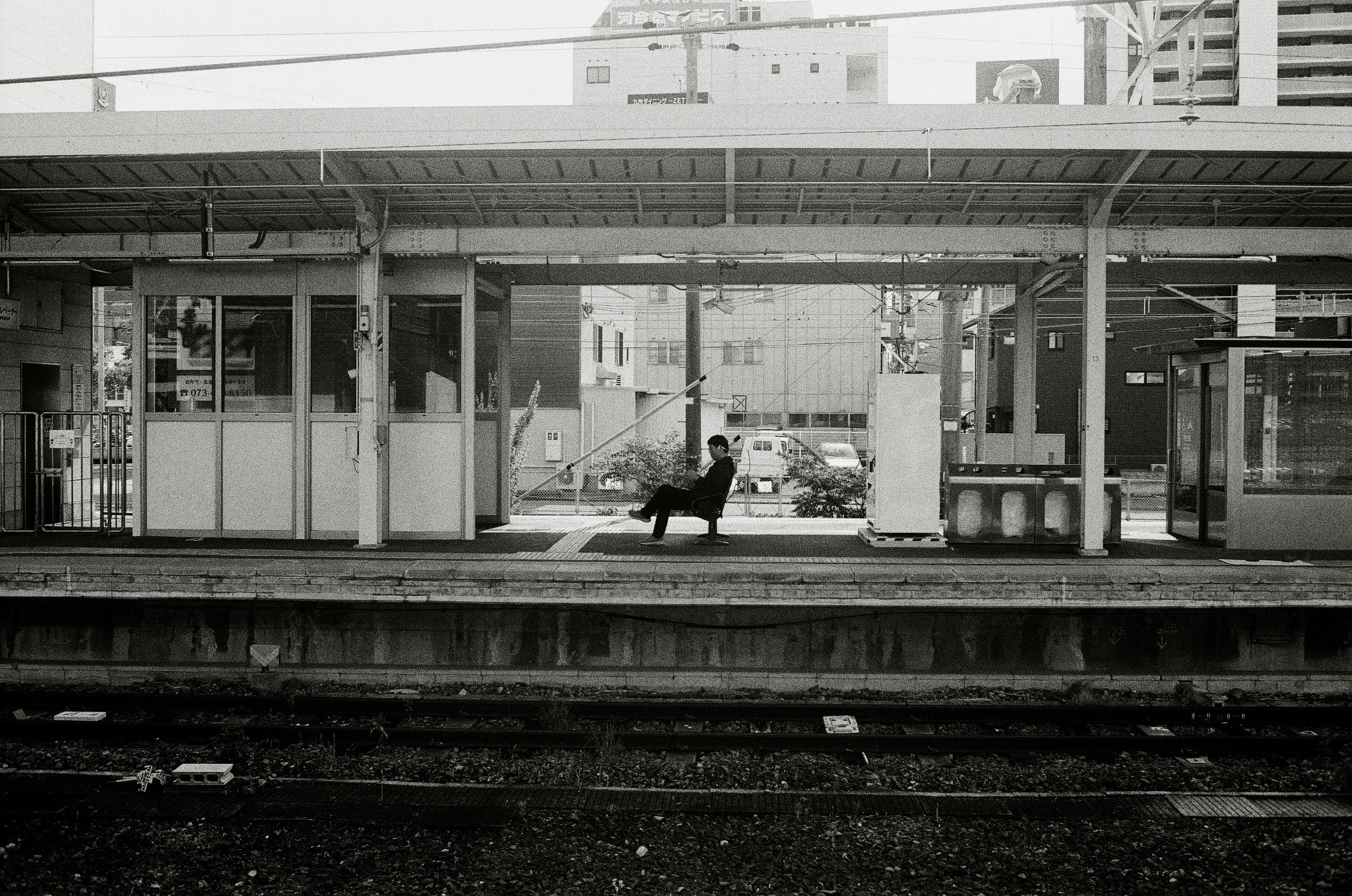 Profile of a man in dark clothes sitting on an bench on an empty train platform. A vending machine is on the right of the frame, and a room of sorts is on the left.