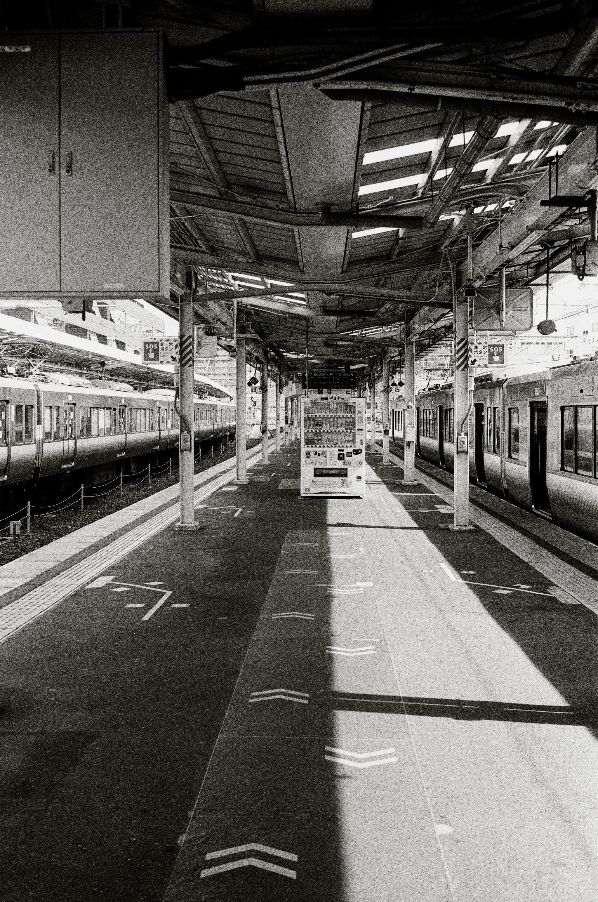 A vending machine is in the centre of the picture, with marked lines on the ground showing arrows pointing to and away from it. The shot is half shaded, with trains on either side of the frame.