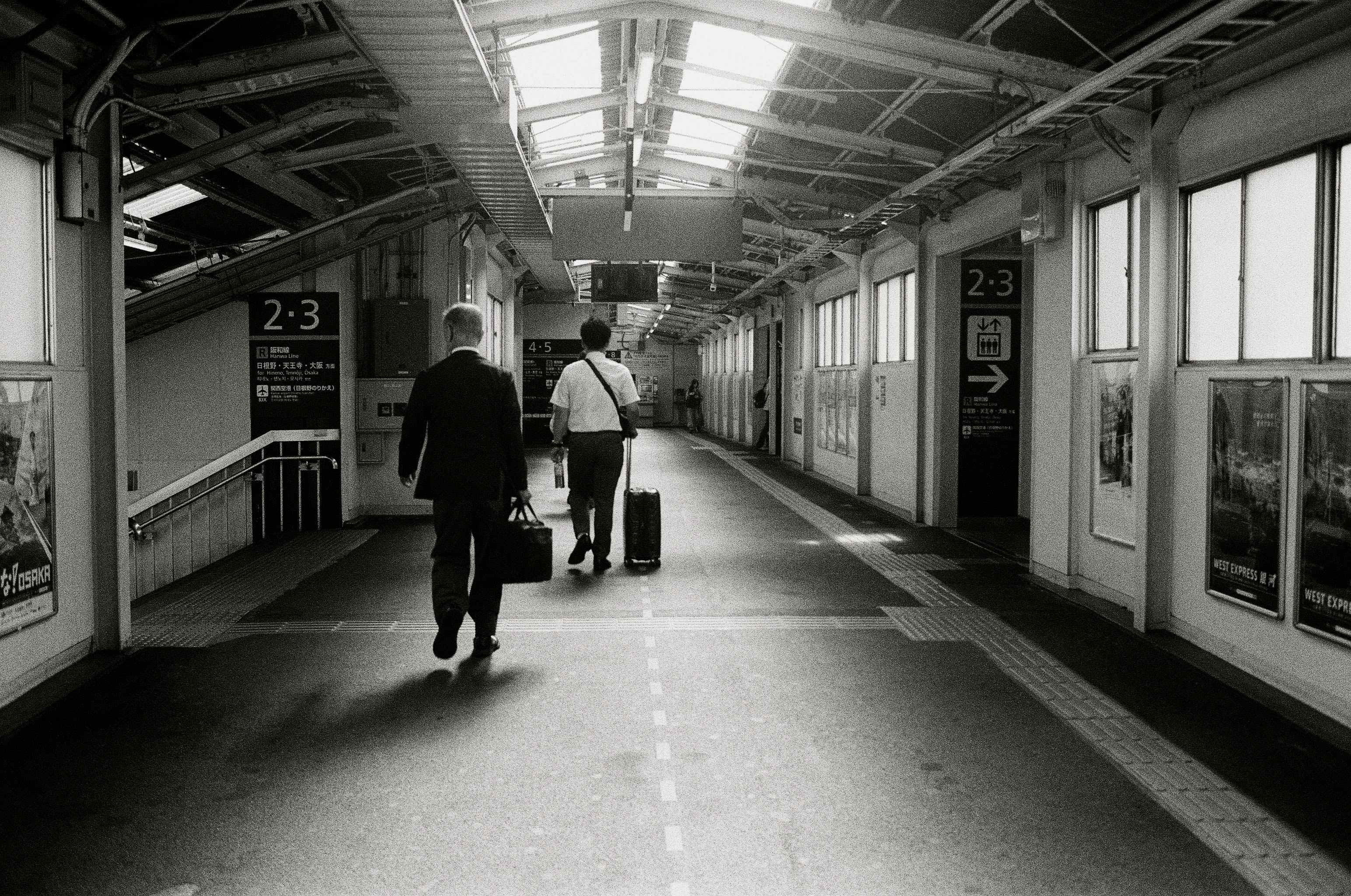 Two business men walking away from the camera on a train station concourse. The man nearest to camera is dressed in a suit and carrying a briefcase. He is looking down a flight of stairs at the platform below. The man furthest from camera wears a white shirt and dark pants, and is pushing a small suitcase.