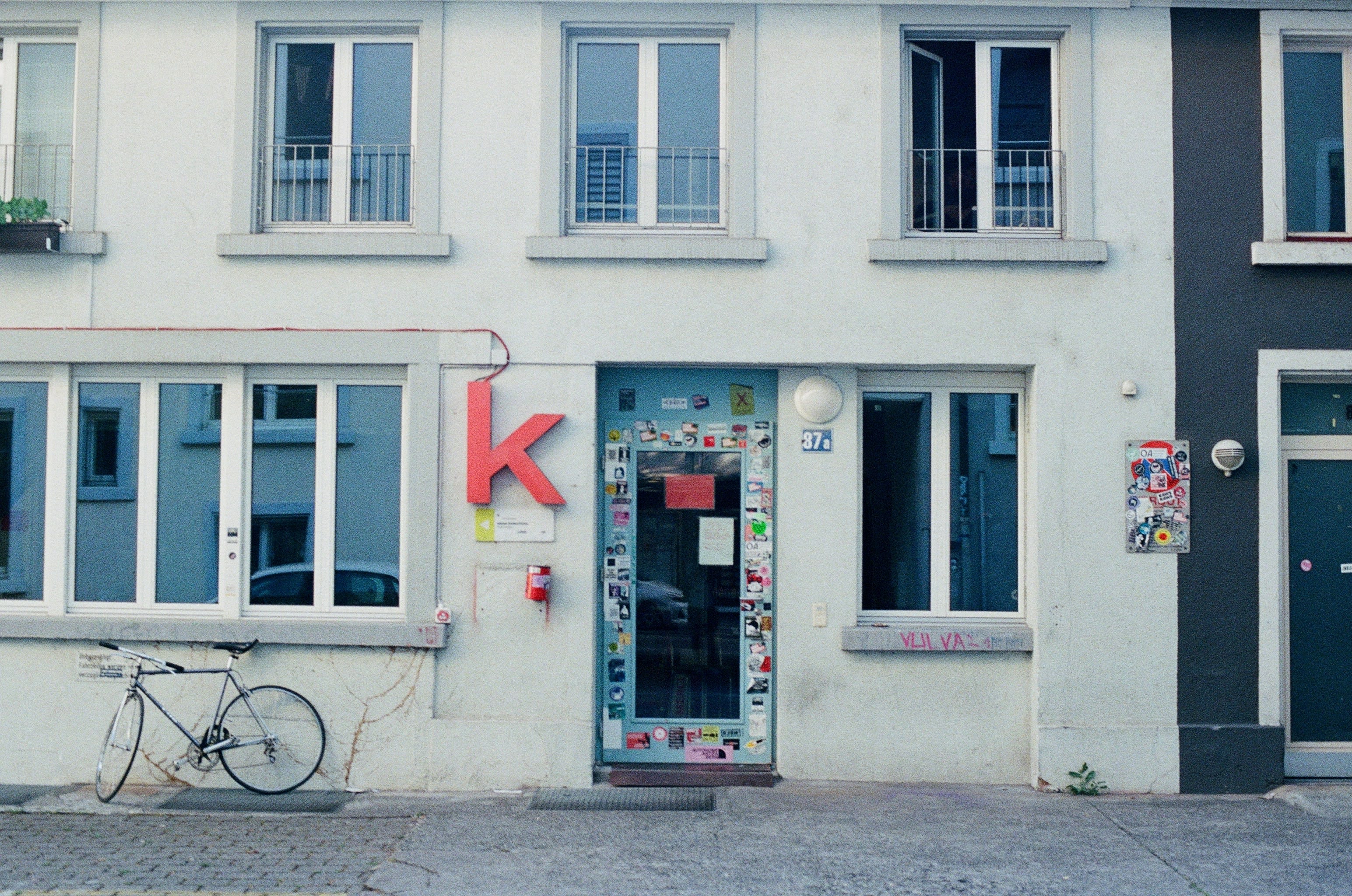 View of the front of a white building. A bike is leaned against it, to the left of a door plastered with stickers. A big red letter K is placed on the left of the doorway.
