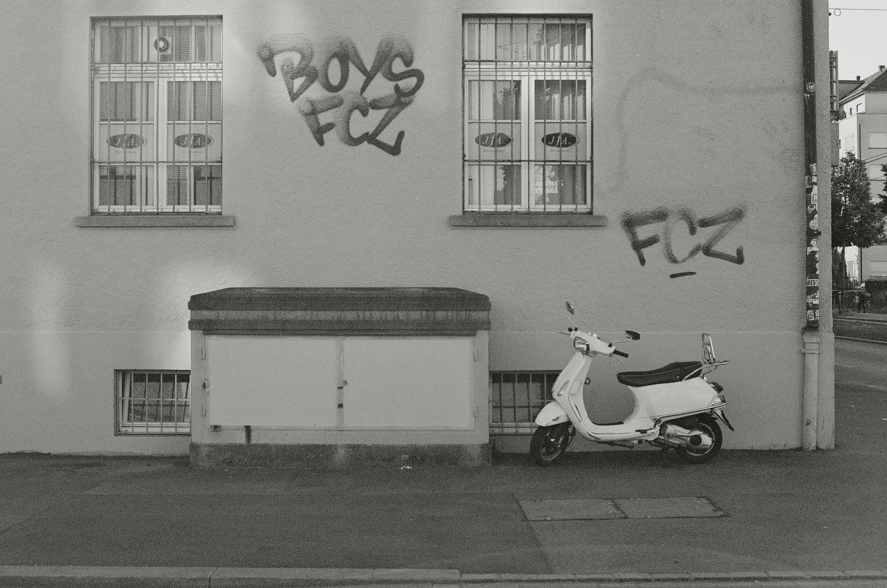 Picture of the side of a building. A scooter is parked to the right of a large garbage bin. Grafitti tags the wall.