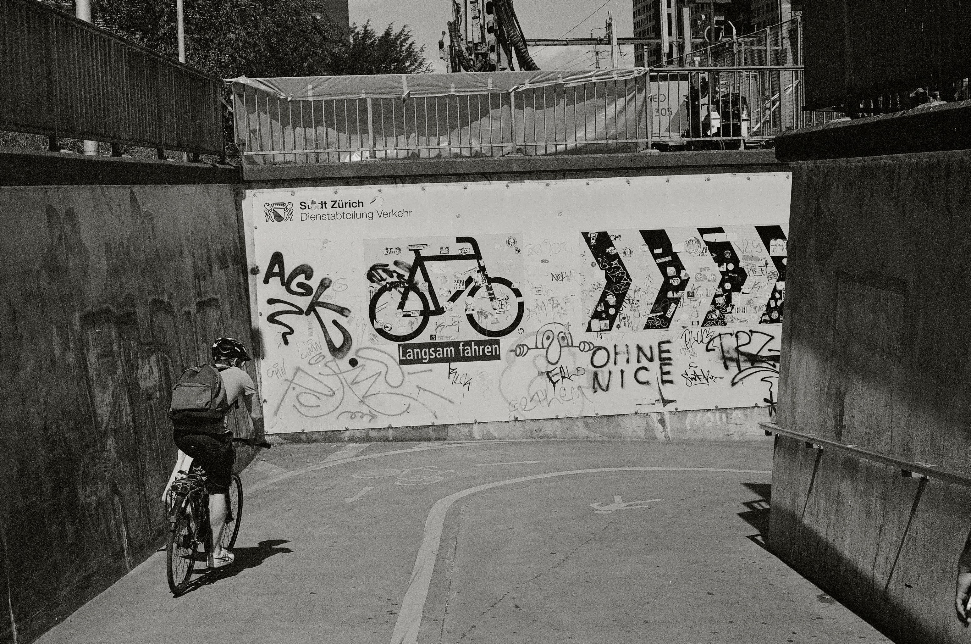 A black and white photo of a cyclisting about to head around a corner into an underpass. Cycling directions are indicated on the walls, alongside large amounts of grafitti.