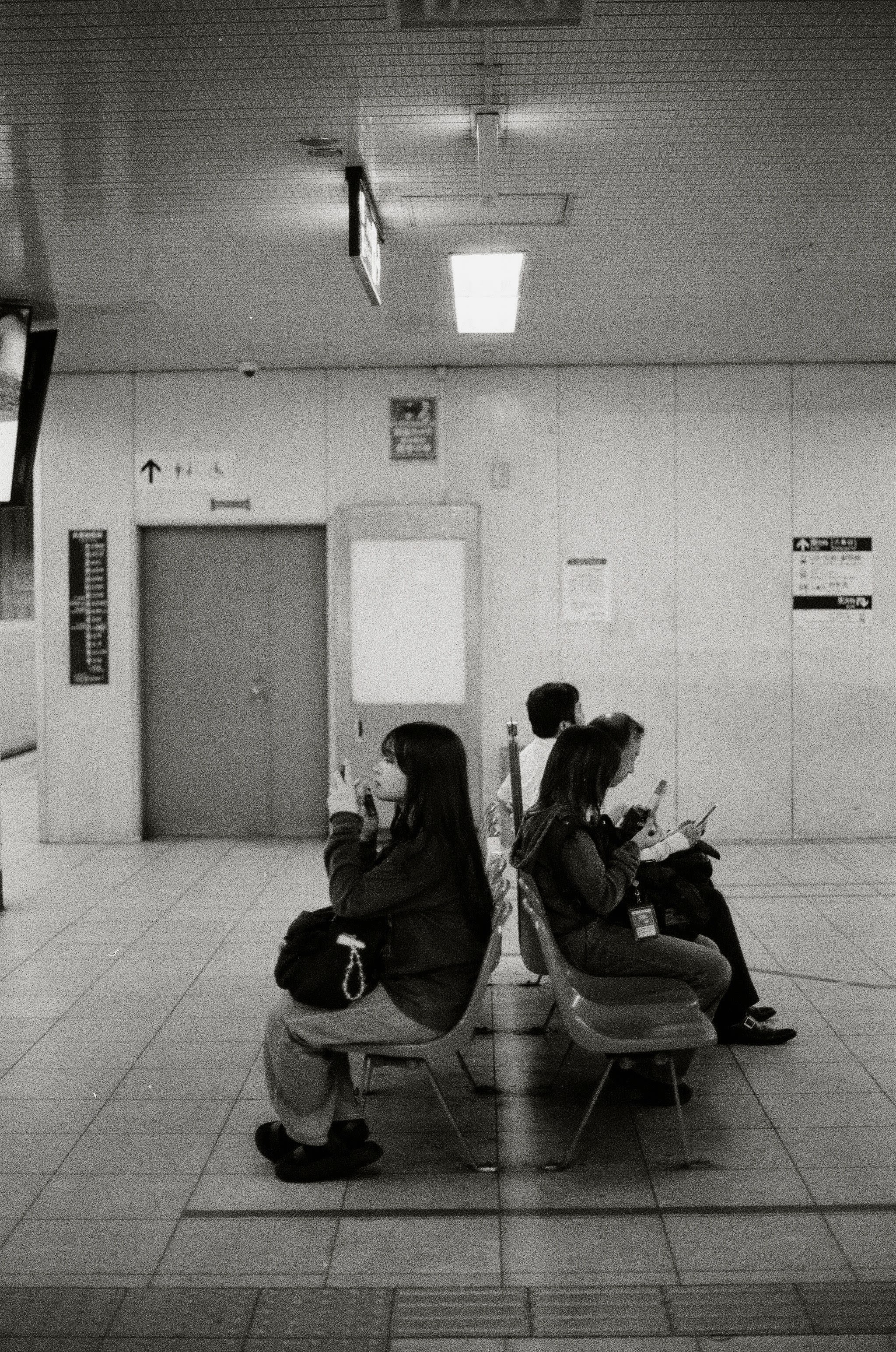Profile of people seated on a train platform, back-to-back. All three people in the shot are looking at their phones.