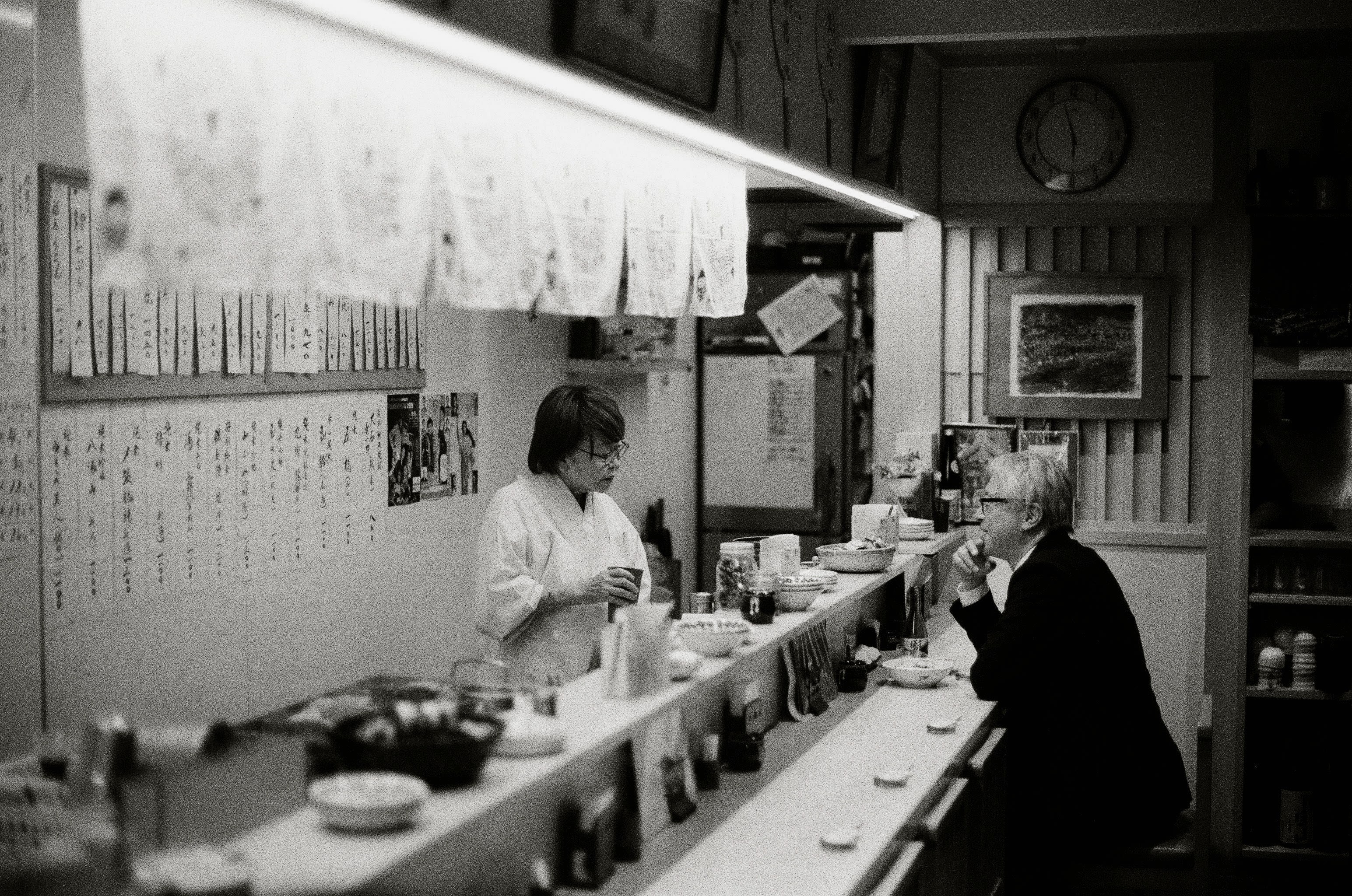 Black and white photo with a seated elderly man in a suit speaking to a woman standing behind the counter.
