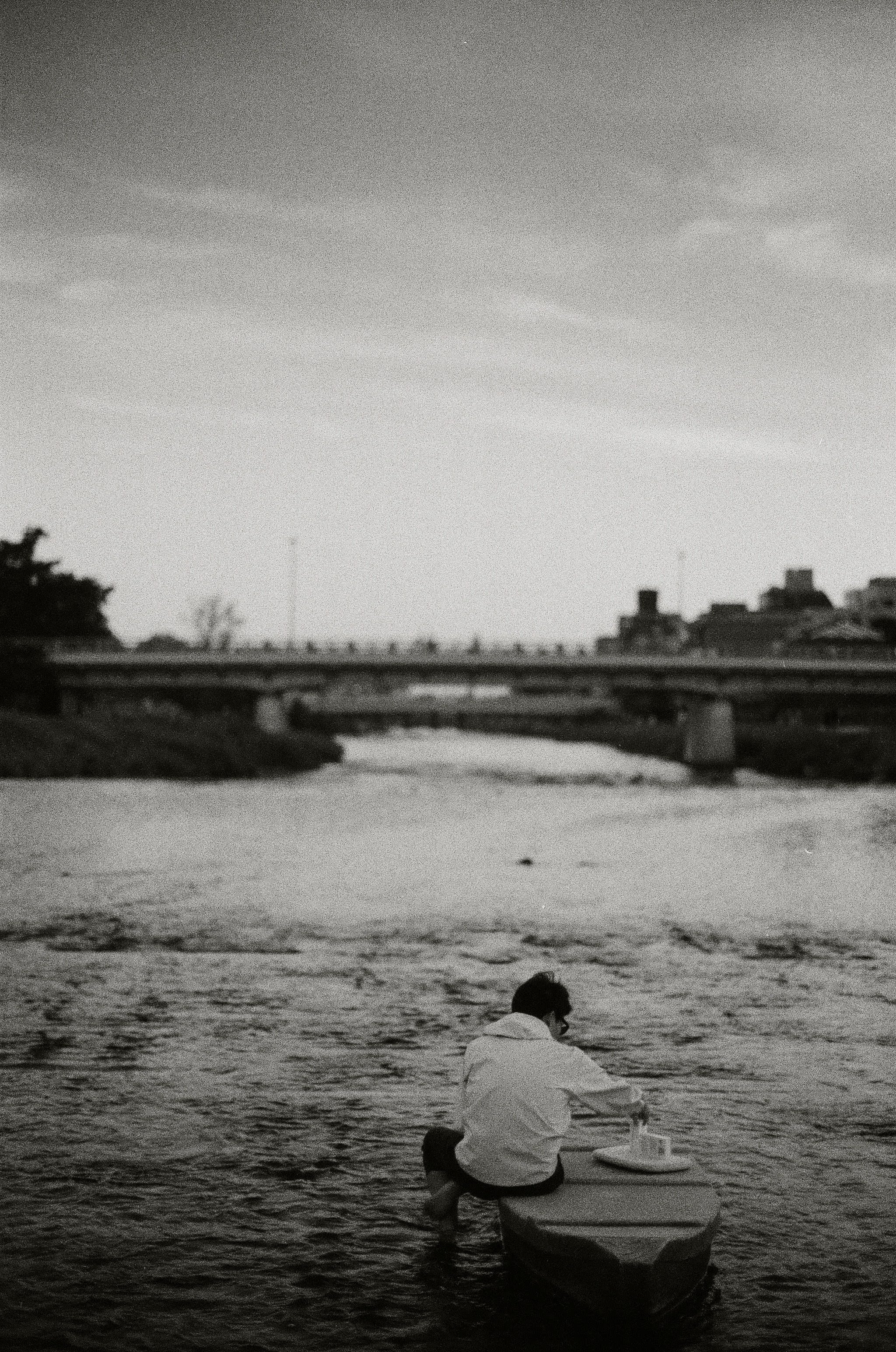 A boy sits on a rock in the middle of a river. The photo is taken from behind the boy, and looks towards a birdge that crosses the river in the midground.