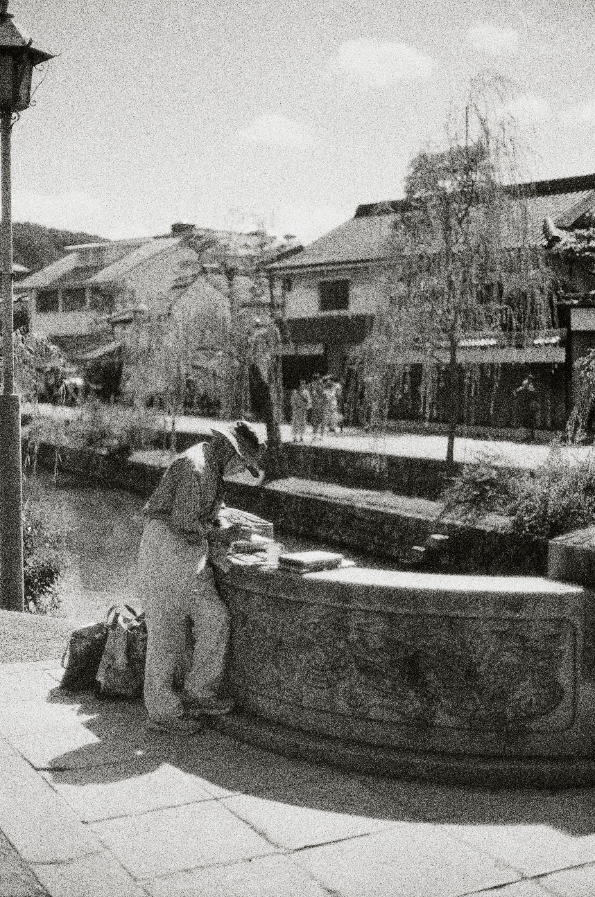 A well dressed elderly man leans on a stone railing, painting the river scene before him.
