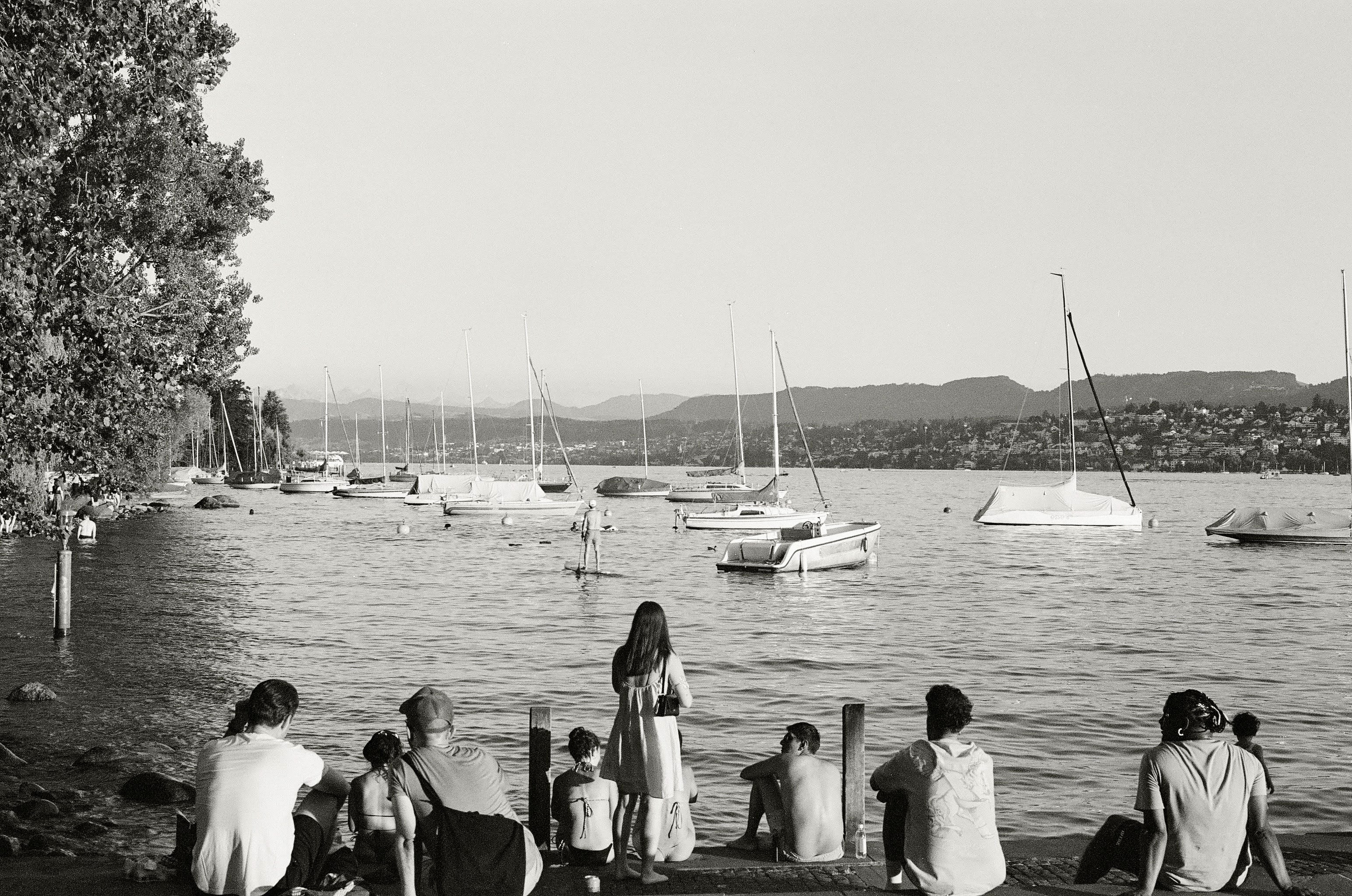 A line of people site by the side of a lake. The photo is taken from behind them, as they look out at the yatchs on the water and mountains in the distance.